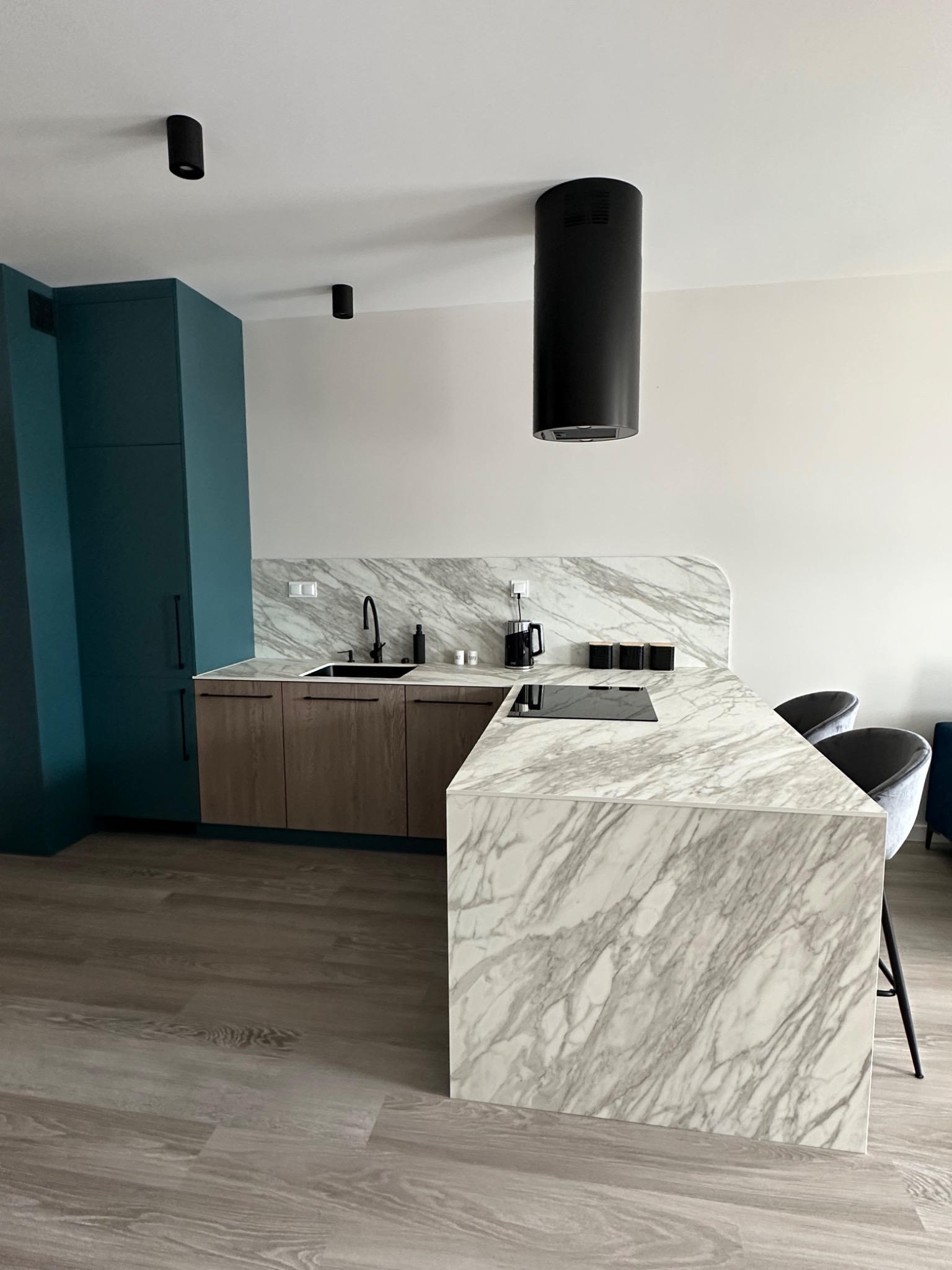 Nowoczesna kuchnia z wyspą wykończoną marmurem, czarne akcenty oświetleniowe i armatura, drewniane szafki dolne, turkusowa zabudowa, panele podłogowe imitujące drewno.