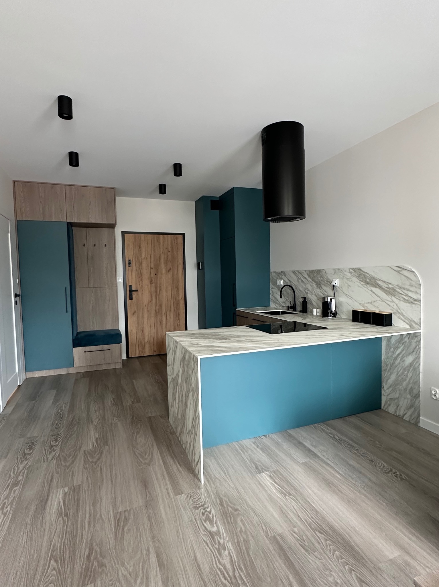 Nowoczesna kuchnia z wyspą, marmurowym blatem i turkusowymi akcentami kolorystycznymi. Widoczne oświetlenie punktowe i drewniana podłoga.