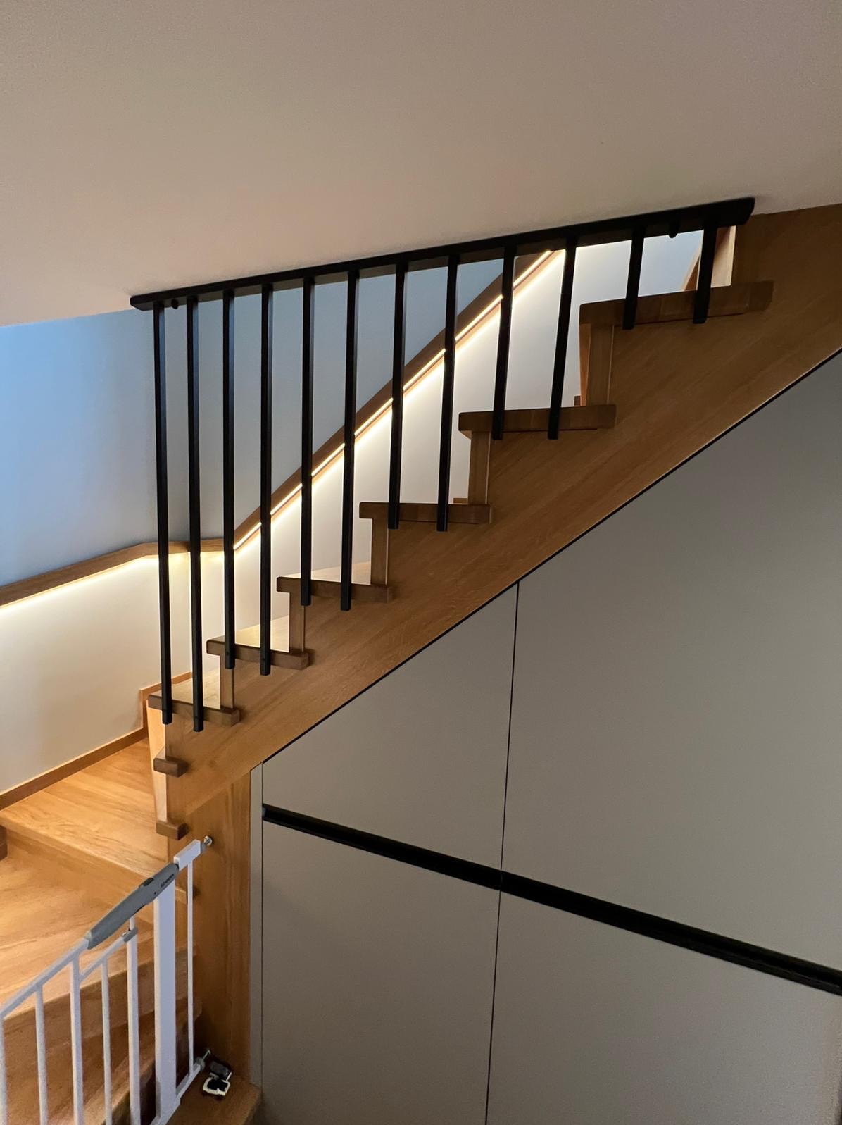 Drewniane schody z czarną balustradą i podświetleniem LED, zintegrowane z zabudową meblową w odcieniach szarości. Widok z góry, nowoczesny design.