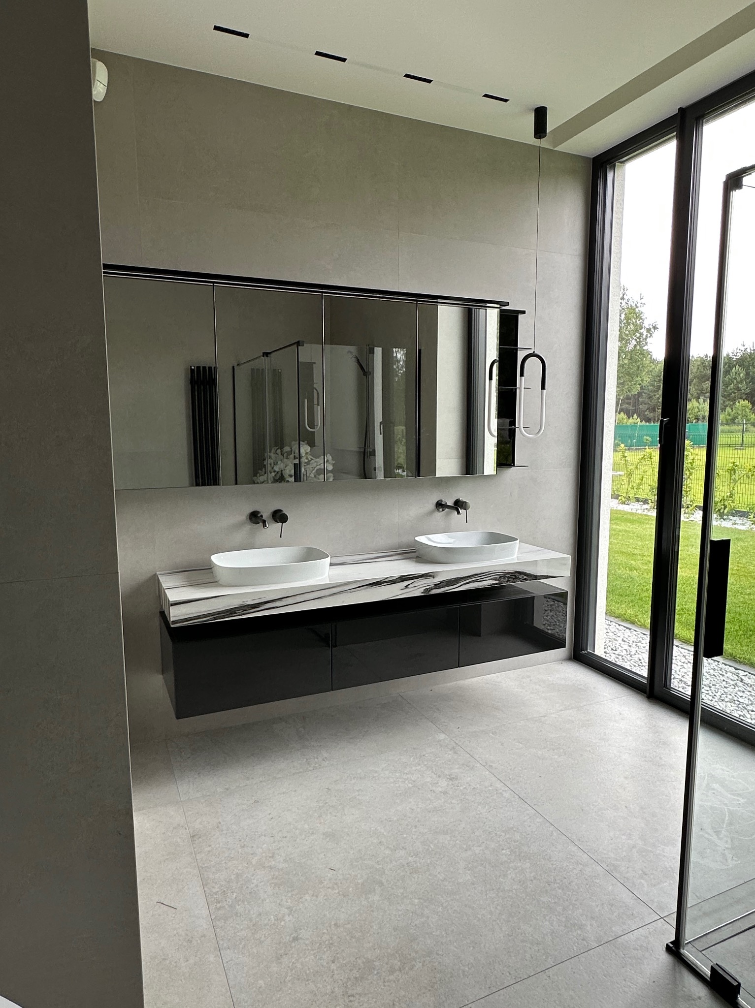 Nowoczesna łazienka z dwoma umywalkami na blacie imitującym marmur, szafką podwieszaną i dużym lustrem szafkowym. Widok na ogród przez panoramiczne okno.