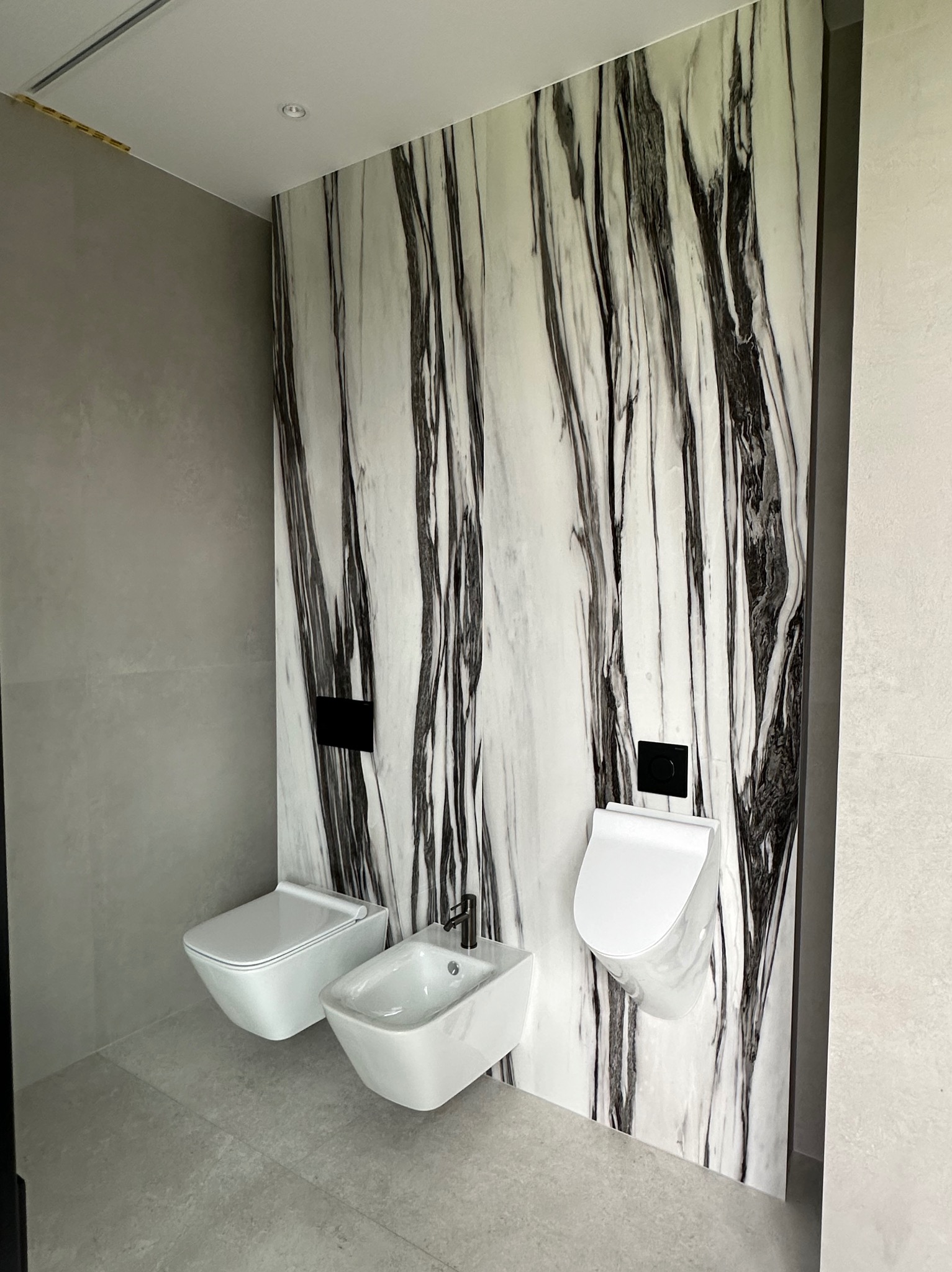 Nowoczesna łazienka z marmurową ścianą akcentującą, podwieszaną toaletą i bidetem. Minimalistyczny design, szare płytki podłogowe, czarne elementy armatury.