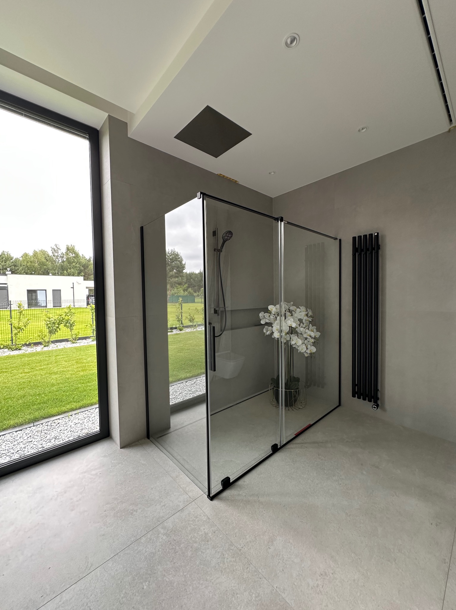 Nowoczesna kabina prysznicowa z czarnymi ramami i kwiatami w jasnej łazience z dużym oknem na ogród. Minimalistyczny design, szare płytki i grzejnik.