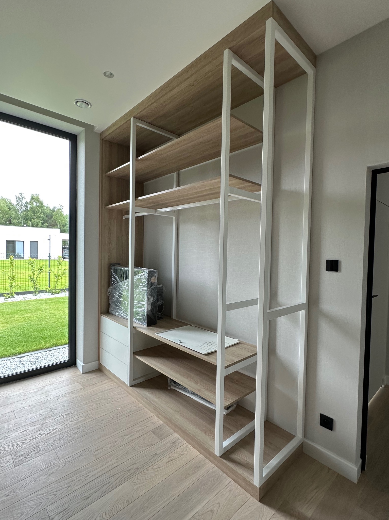 Nowoczesna zabudowa meblowa z jasnego drewna i białych elementów metalowych, w jasnym wnętrzu z widokiem na ogród przez okno. Minimalistyczny design.