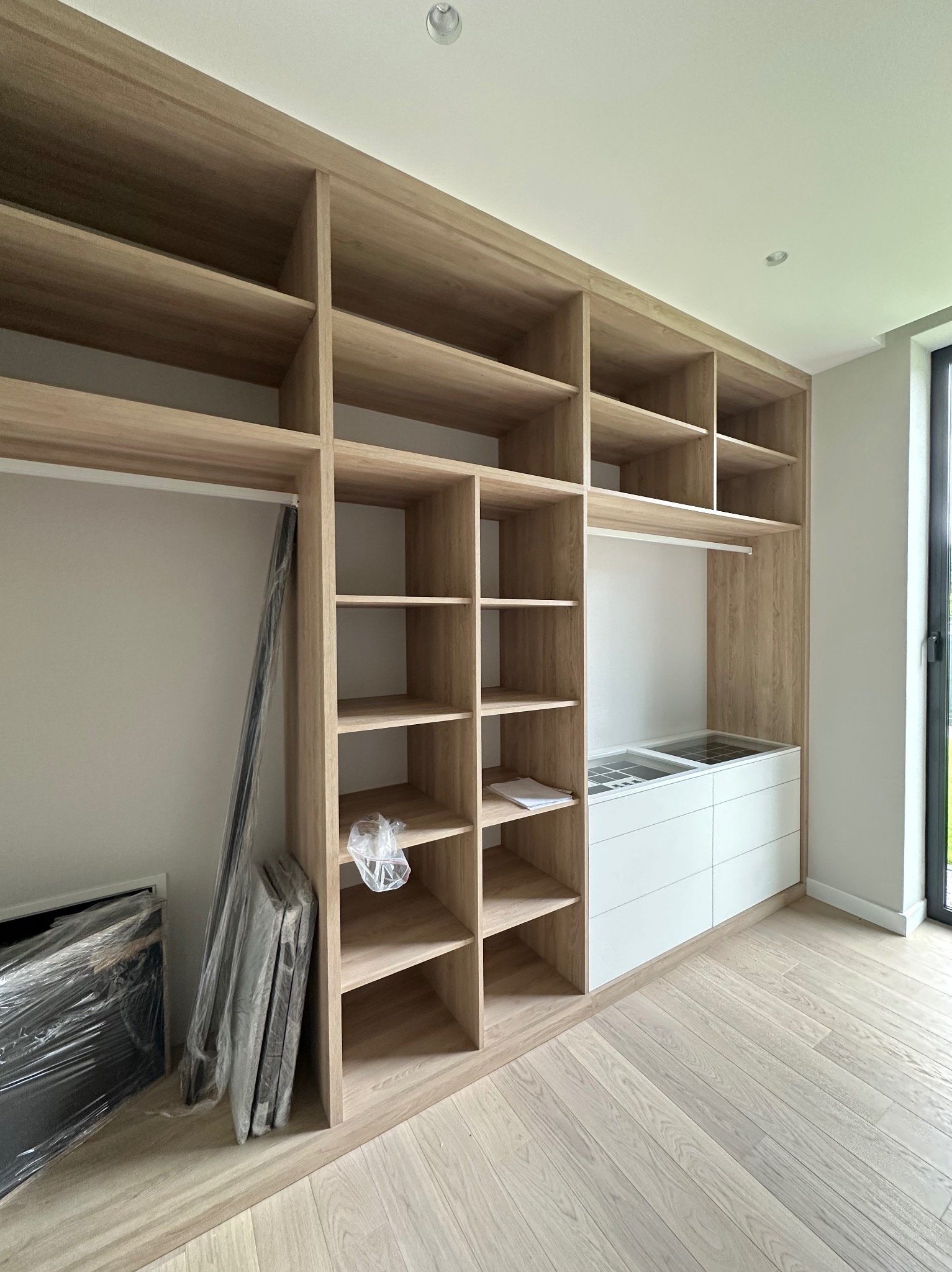 Jasne drewniane regały z półkami i białymi szufladami tworzą przestrzeń garderoby. Widoczne elementy w folii ochronnej, podłoga z jasnego drewna. Nowoczesne wykończenie.