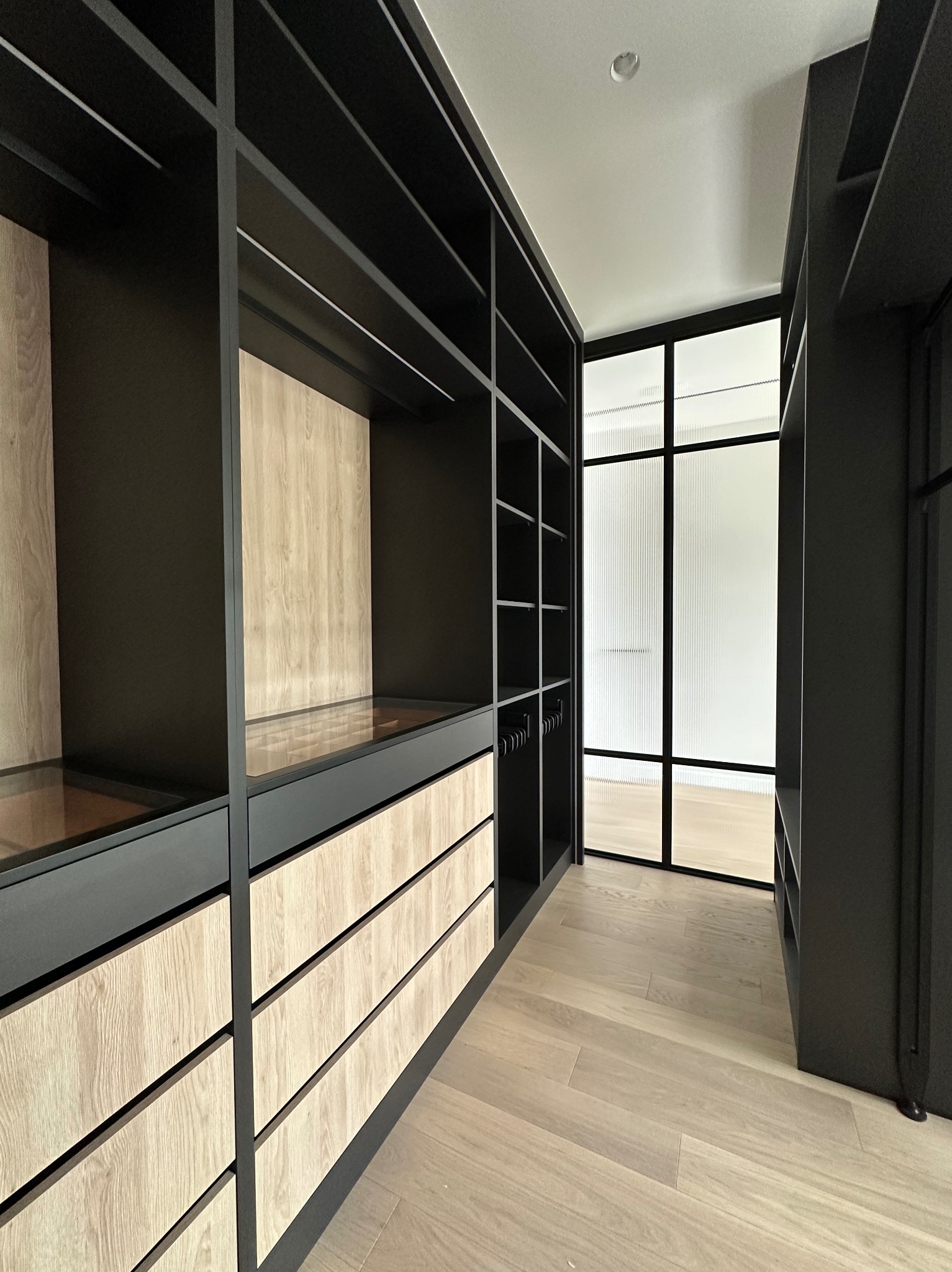 Nowoczesna garderoba z czarnymi ramami i drewnianymi frontami szuflad, widok na przeszklone drzwi. Jasna podłoga z paneli.