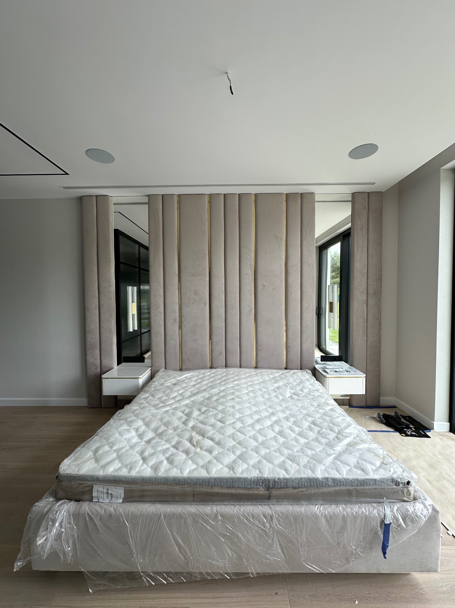 Beżowe łóżko z tapicerowanym wezgłowiem z pionowymi panelami i złotymi akcentami. Na szafkach nocnych lustra. Całość w jasnym, nowoczesnym wnętrzu.