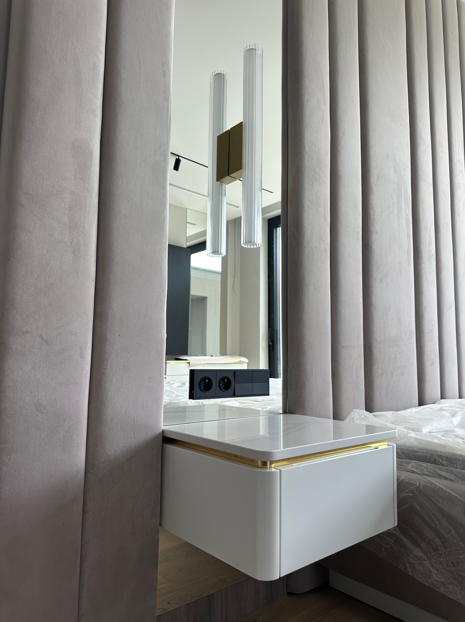 Elegancka sypialnia z panelem ściennym, szafką nocną z marmurowym blatem i złotymi akcentami. Widoczne gniazdka elektryczne i lampy wiszące. Nowoczesny design.