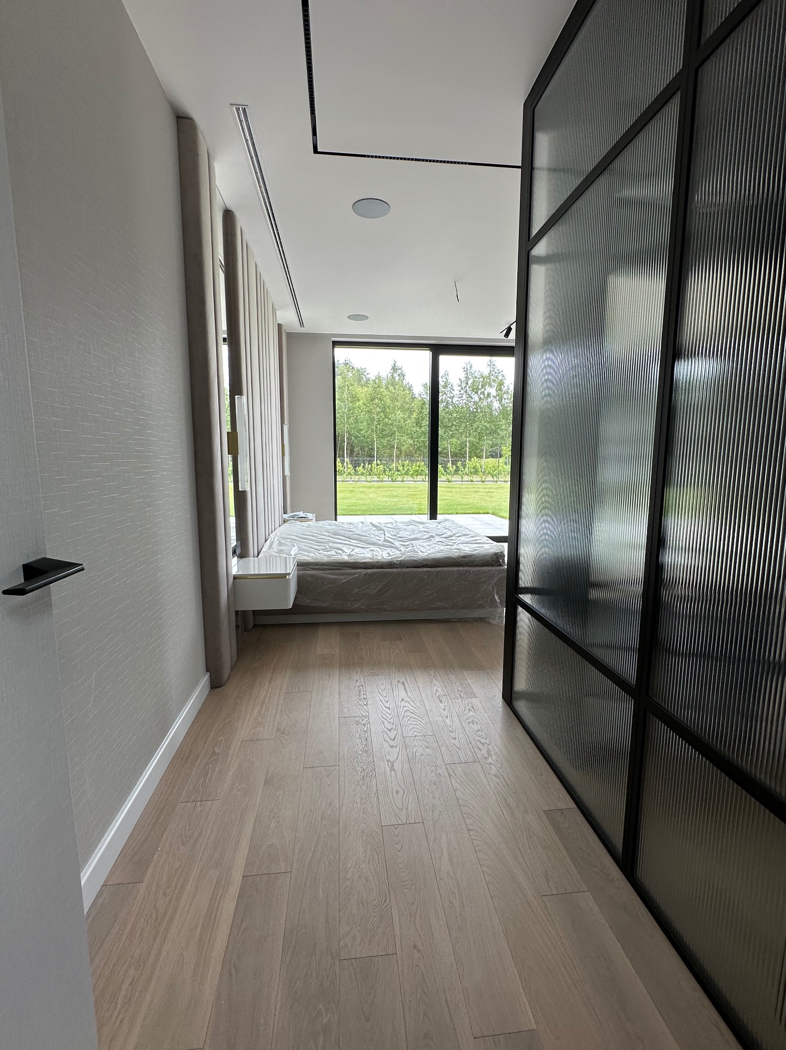 Nowoczesna sypialnia z parkietem, szklaną ścianą działową i widokiem na zieleń za oknem. Minimalistyczny design z elementami tapicerowanymi i eleganckim oświetleniem.