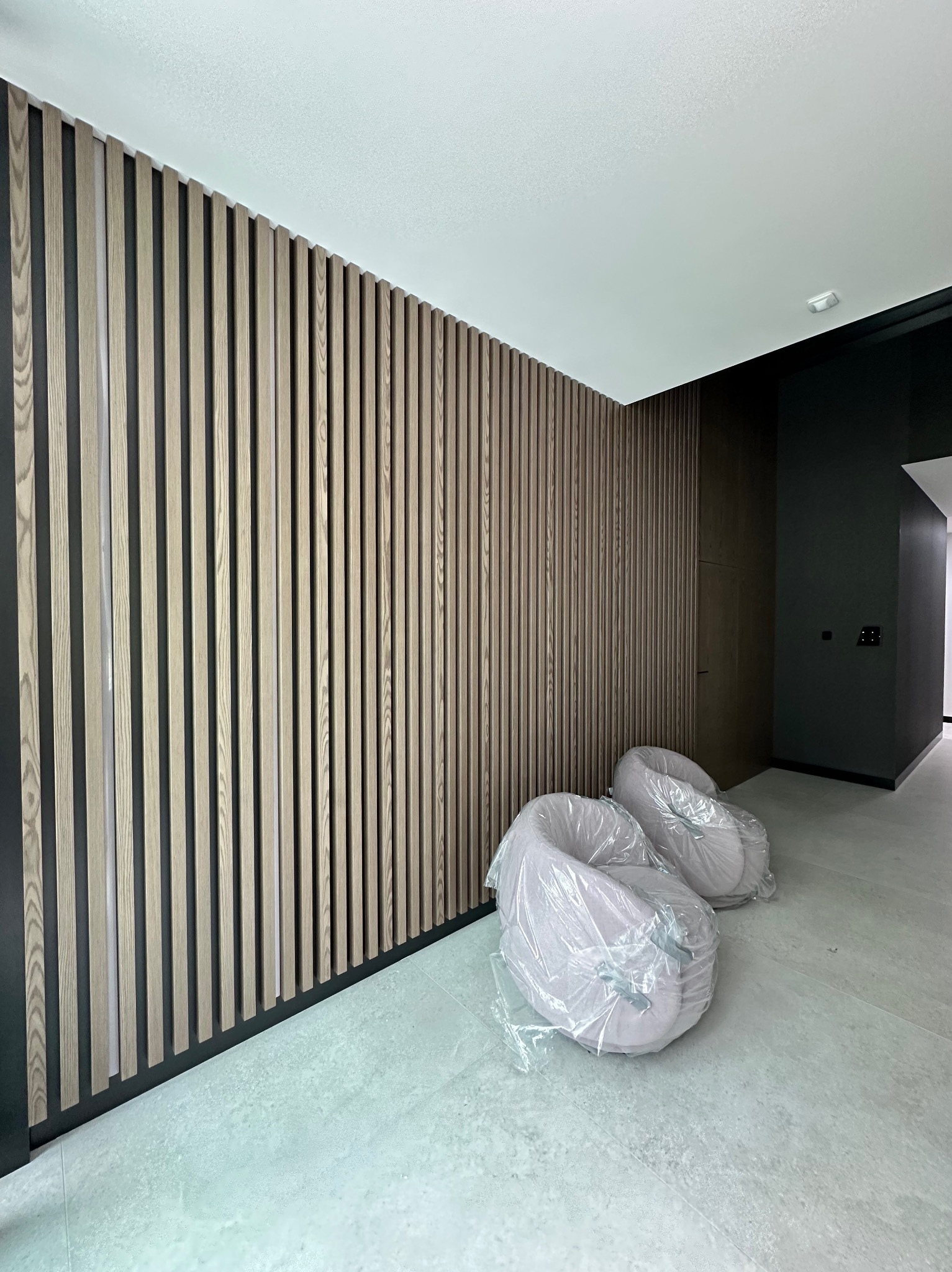 Drewniana ściana lamelowa w nowoczesnym wnętrzu, z dwoma fotelami w folii ochronnej. Minimalistyczny design z czarnymi akcentami i jasną podłogą.