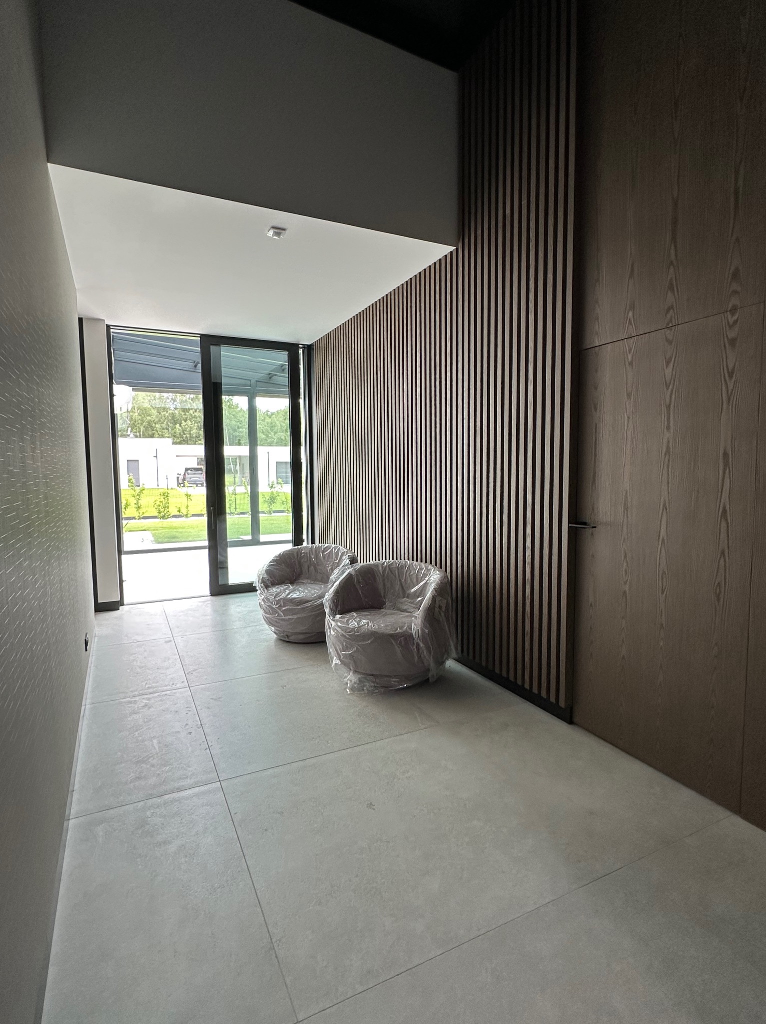 Nowoczesny hol z drewnianą ścianą z lameli, szarą podłogą z płytek i dwoma fotelami okrytymi folią ochronną. Minimalistyczny design wnętrza.