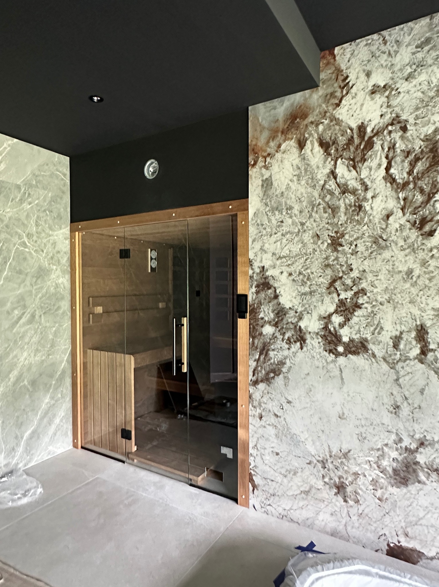 Nowoczesna sauna z przeszklonymi drzwiami i drewnianym wykończeniem, na tle ściany z efektem marmuru i ciemnego sufitu. Całość w jasnej, minimalistycznej kolorystyce.