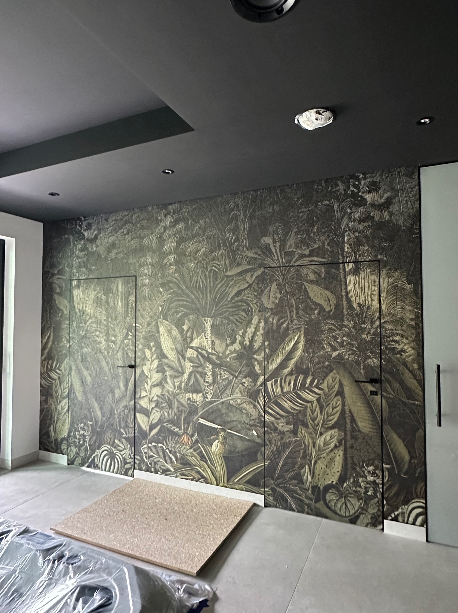 Wnętrze z fototapetą w motywy roślinne, imitującą dżunglę, z ukrytymi drzwiami i czarnym sufitem. Na podłodze płyta i folia ochronna.
