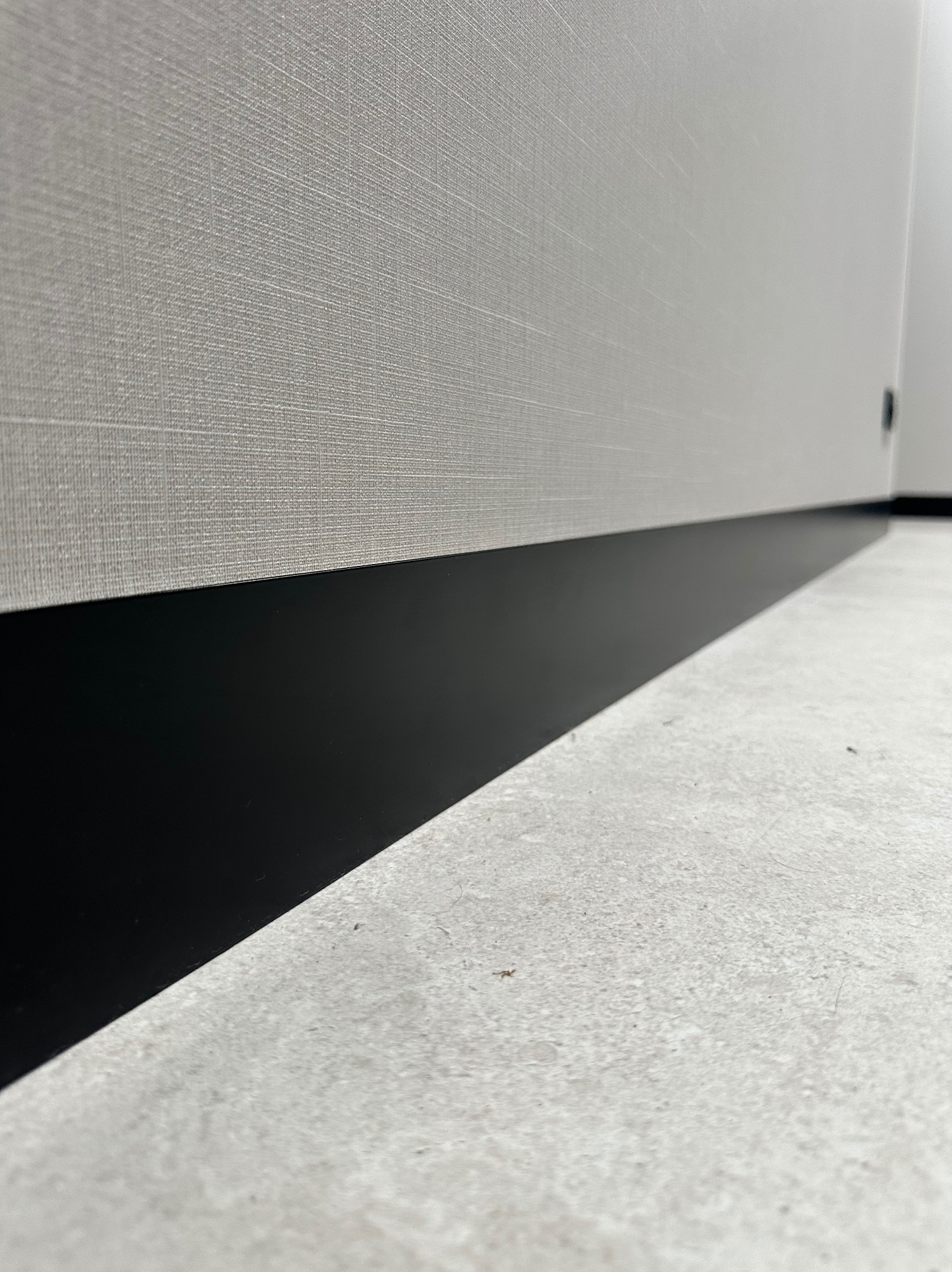 Ściana wykończona tkaniną z czarną listwą przypodłogową i szarą posadzką. Minimalistyczne wnętrze z bliska, ukazujące detale wykończenia.