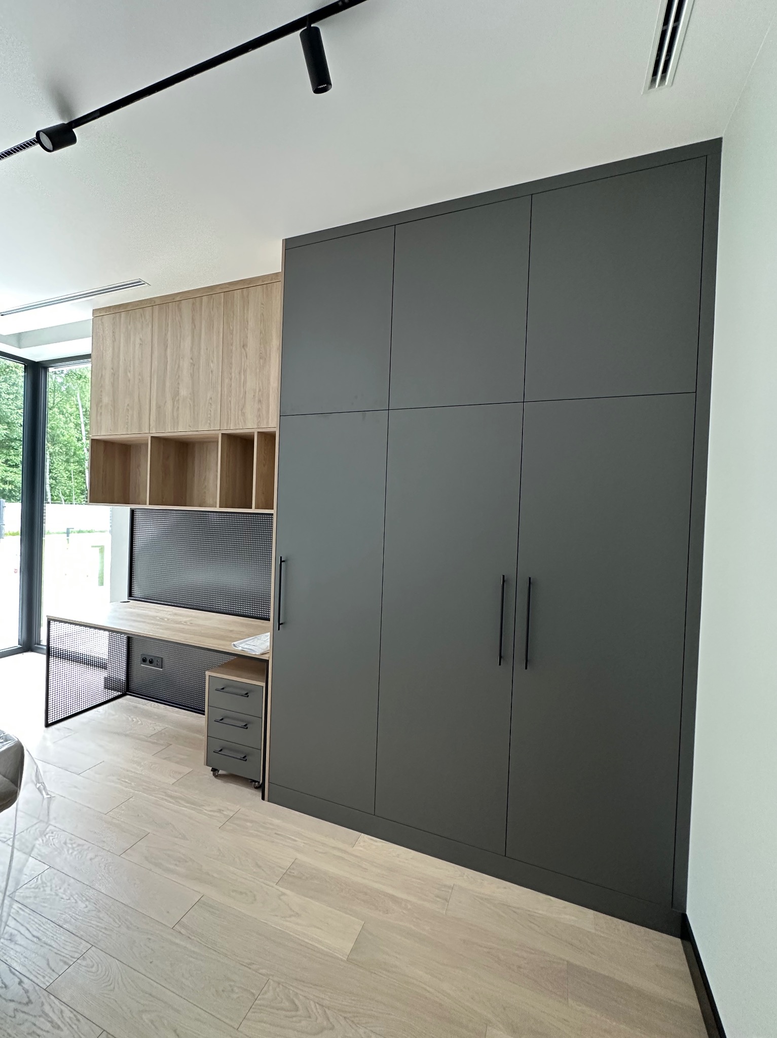 Nowoczesny pokój z zabudową meblową: szara szafa z długimi uchwytami, biurko z drewnianym blatem i metalową siatką, drewniane szafki wiszące. Oświetlenie szynowe.