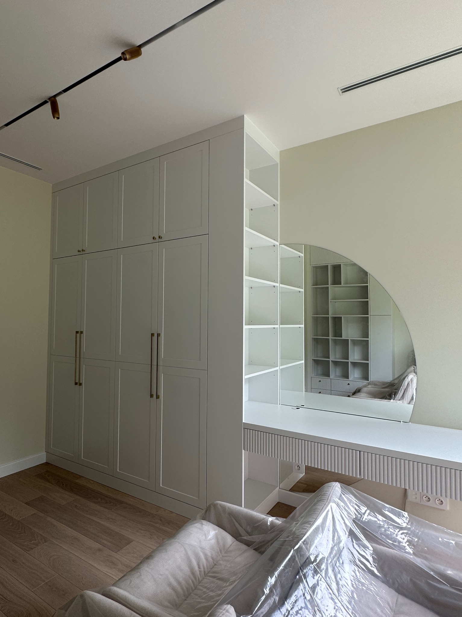 Biała zabudowa meblowa z szafą i toaletką z okrągłym lustrem, uzupełniona oświetleniem szynowym na suficie. Na podłodze panele. Część mebli zabezpieczona folią.