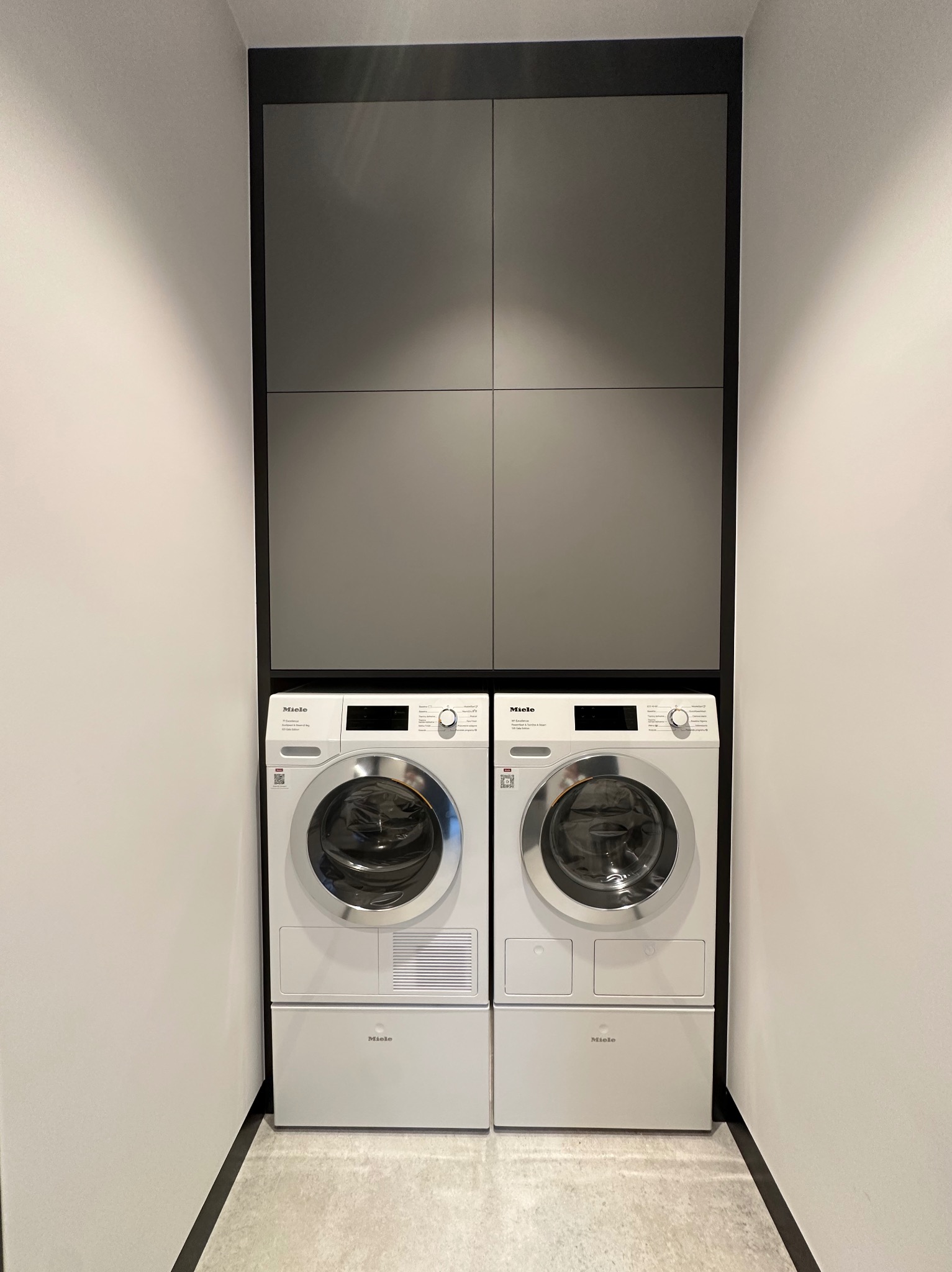 Nowoczesna zabudowa pralni z szarymi szafkami i dwoma pralkami Miele w minimalistycznym wnętrzu. Czyste linie i stonowana kolorystyka.