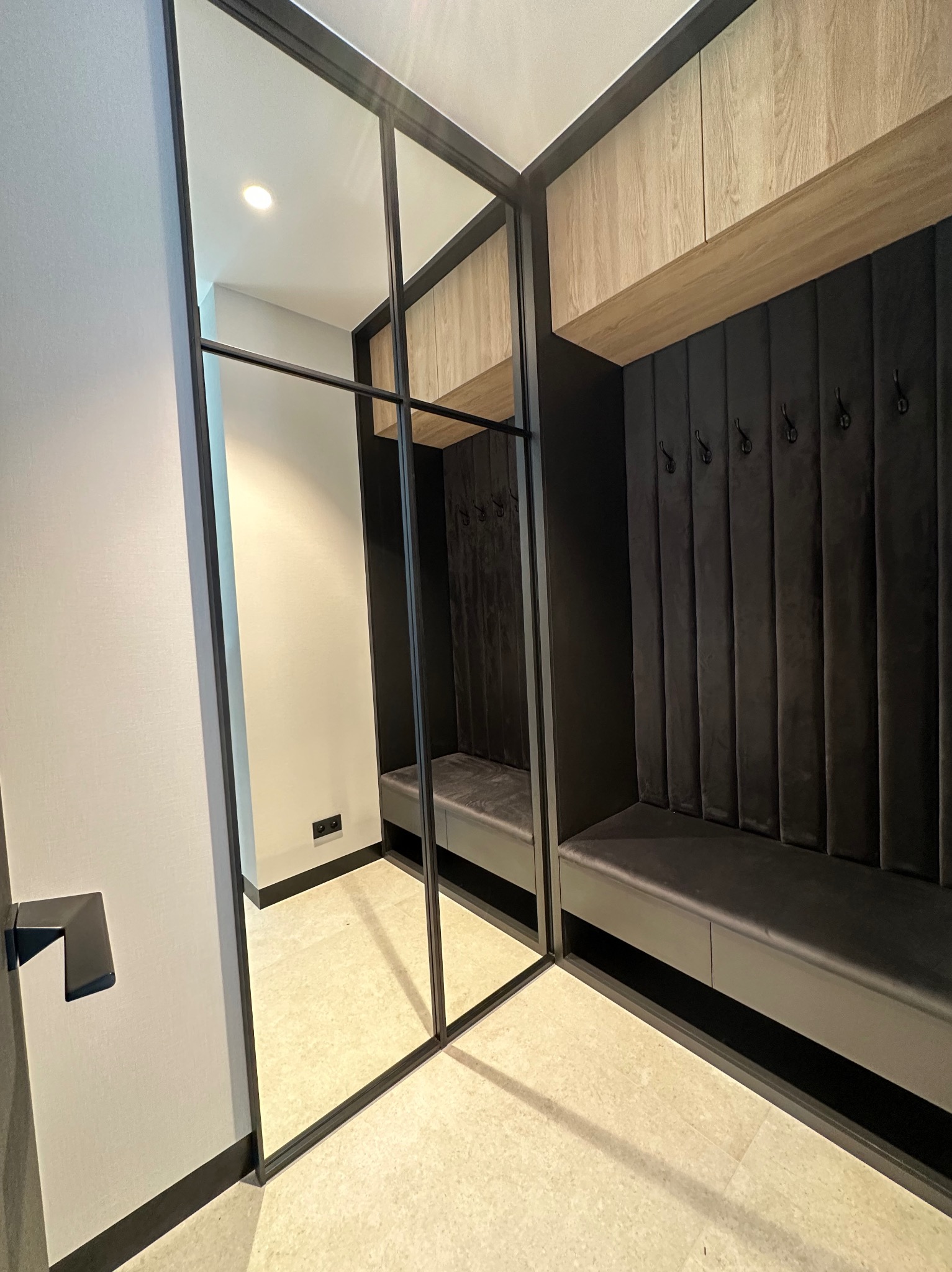 Nowoczesna garderoba w przedpokoju: zabudowa z siedziskiem, wieszakami i szafkami. Lustrzane drzwi przesuwne z czarnymi profilami. Stonowana kolorystyka, eleganckie wykończenie.