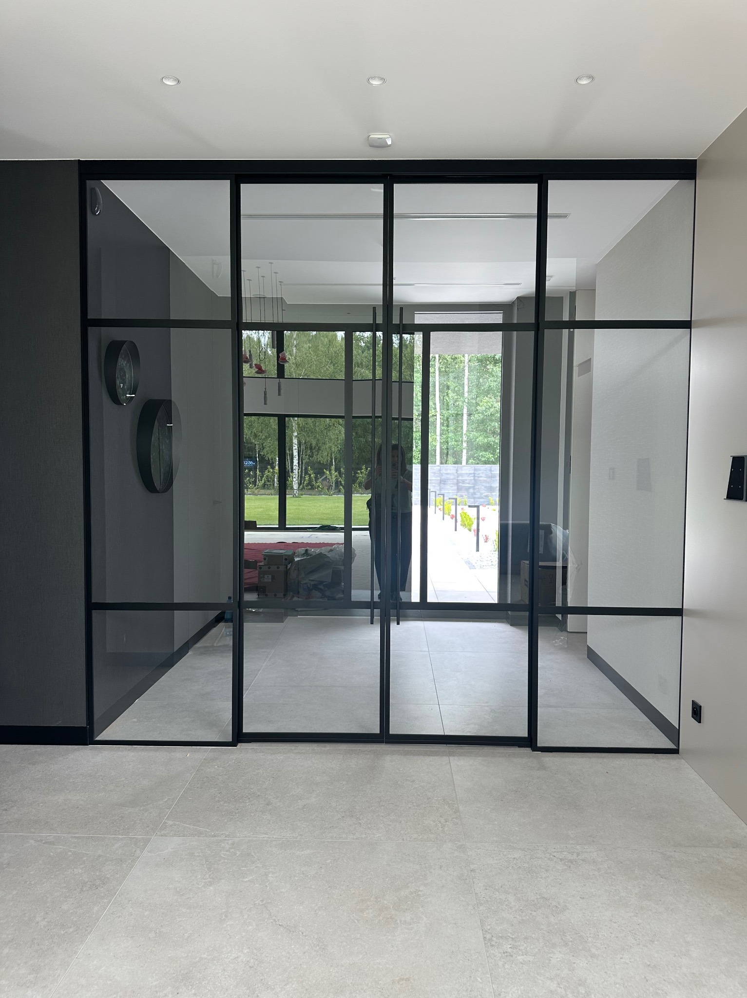 Nowoczesne, czarne, przeszklone drzwi przesuwne oddzielające wnętrze od ogrodu. Widoczne dwa okrągłe zegary na ścianie. Jasna podłoga.