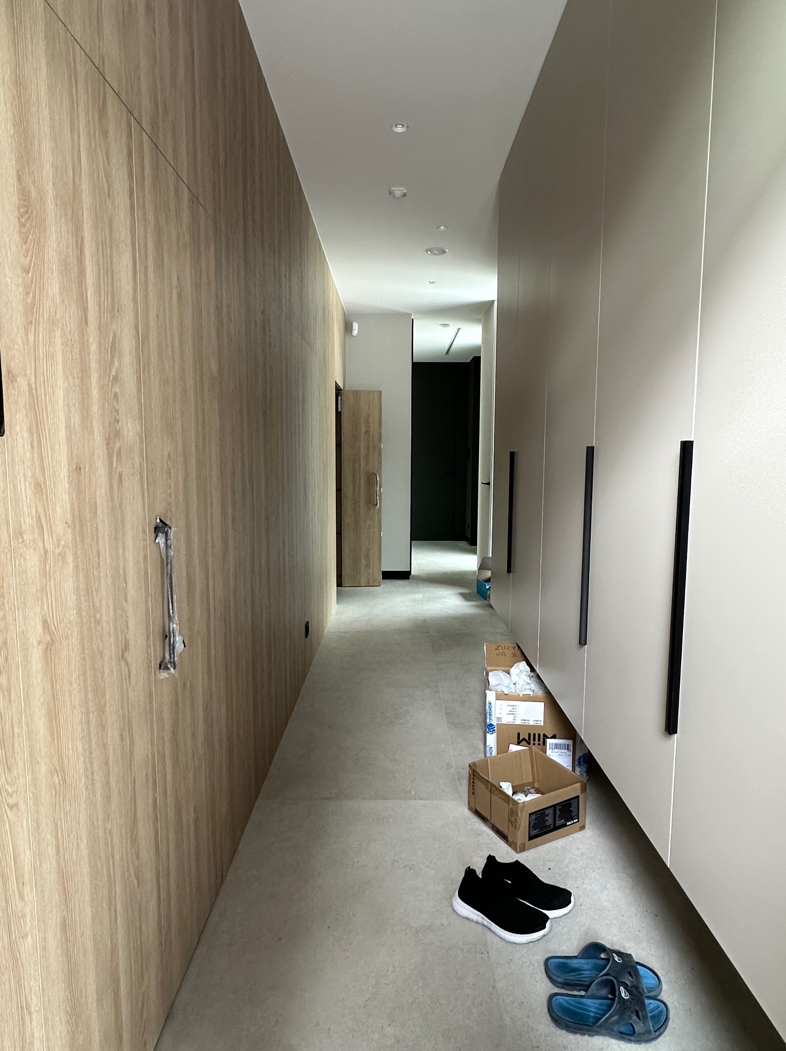 Długi korytarz z zabudową meblową po obu stronach. Po lewej stronie ściana wykończona drewnopodobnymi panelami, po prawej szafy w jasnym kolorze z czarnymi uchwytami.