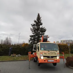 Żółto-pomarańczowy samochód z wysięgnikiem na parkingu, ozdobiona choinka w tle, pracownik w oddali przy dekoracjach.