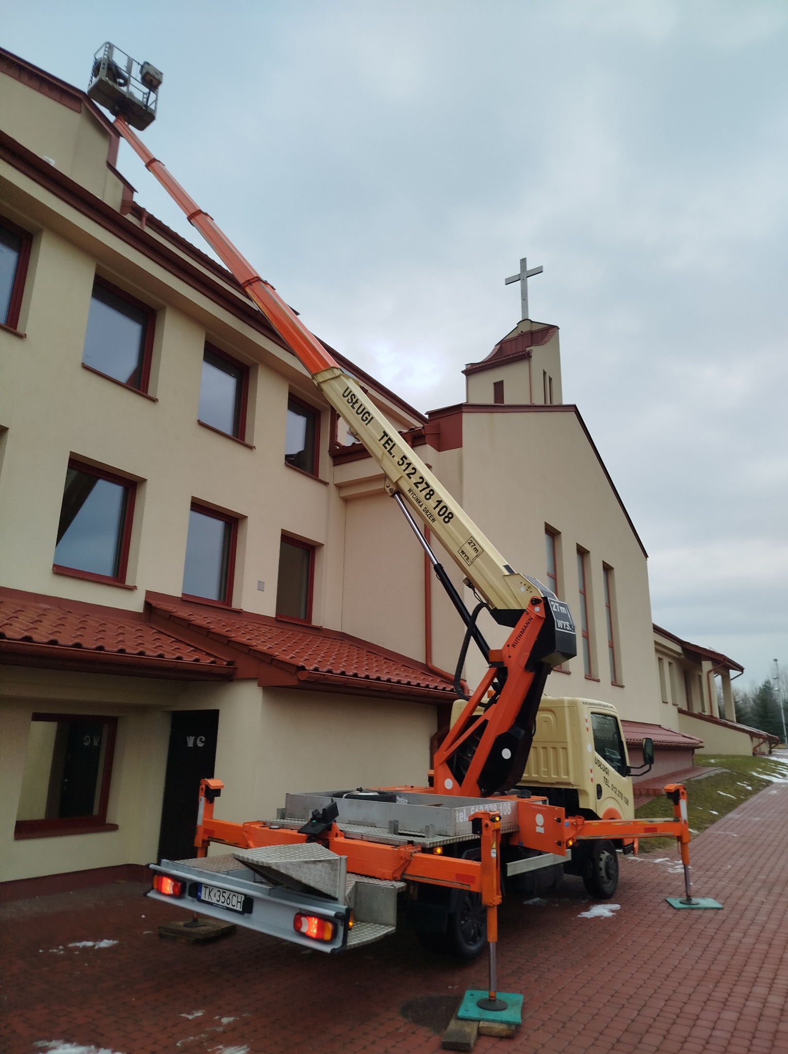 Pomarańczowy podnośnik koszowy na tle budynku kościoła z krzyżem na szczycie, wysięgnik podniesiony do góry, tablica na wysięgniku z napisem 'Wycinka drzew' i numerem telefonu.