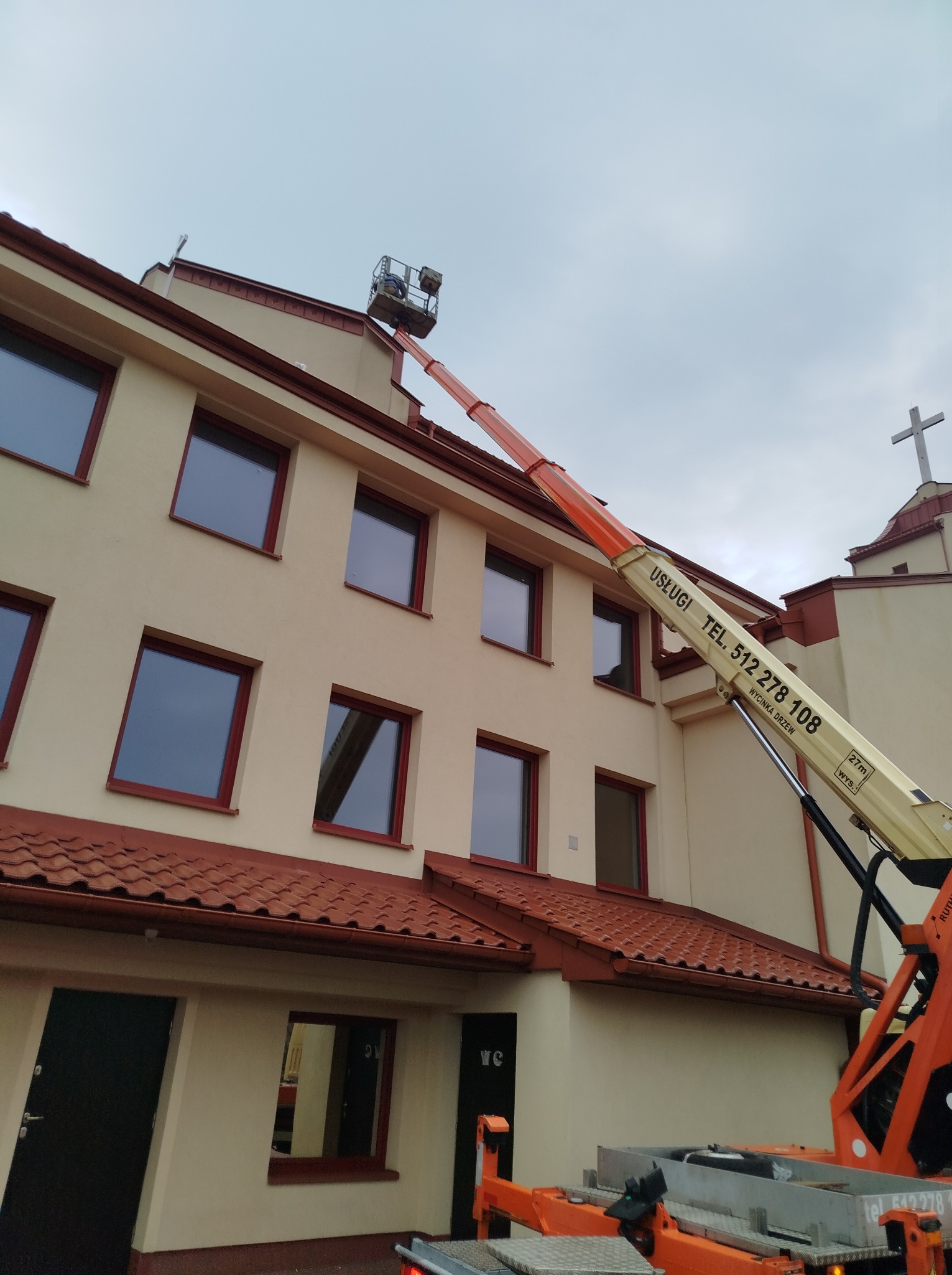 Pomarańczowy podnośnik koszowy z operatorem przy budynku kościoła, prawdopodobnie podczas prac związanych z wycinką drzew, na ramieniu podnośnika widoczny numer telefonu i napis 'Wycinka Drzew'.