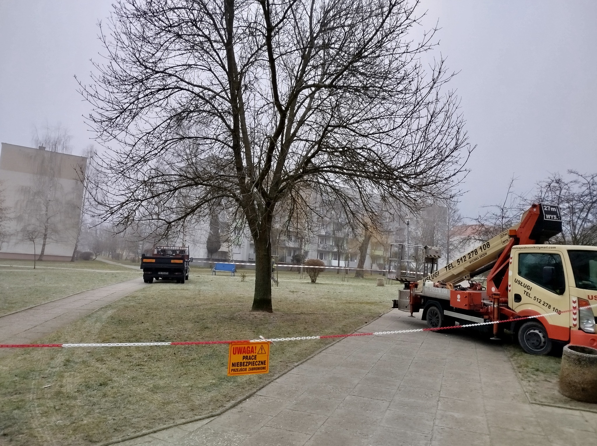 Usługa wycinki drzew w trakcie realizacji: Podnośnik koszowy z wysięgnikiem i ciężarówka na tle drzewa bez liści, teren zabezpieczony taśmą ostrzegawczą i znakiem informującym o niebezpiecznych...