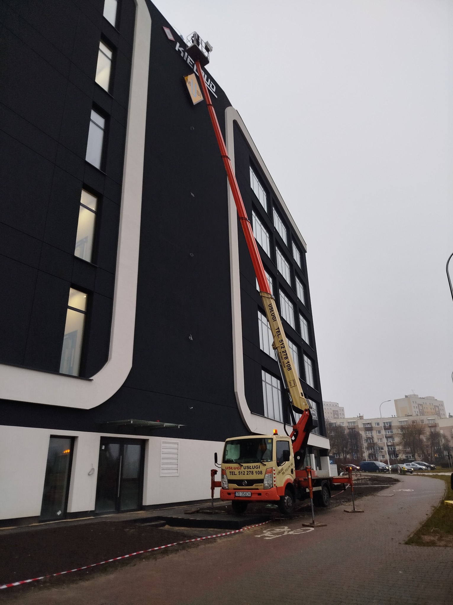 Wysięgnik z platformą podnosi pracownika do montażu lub naprawy litery na elewacji nowoczesnego budynku biurowego o czarnej fasadzie z białymi akcentami.