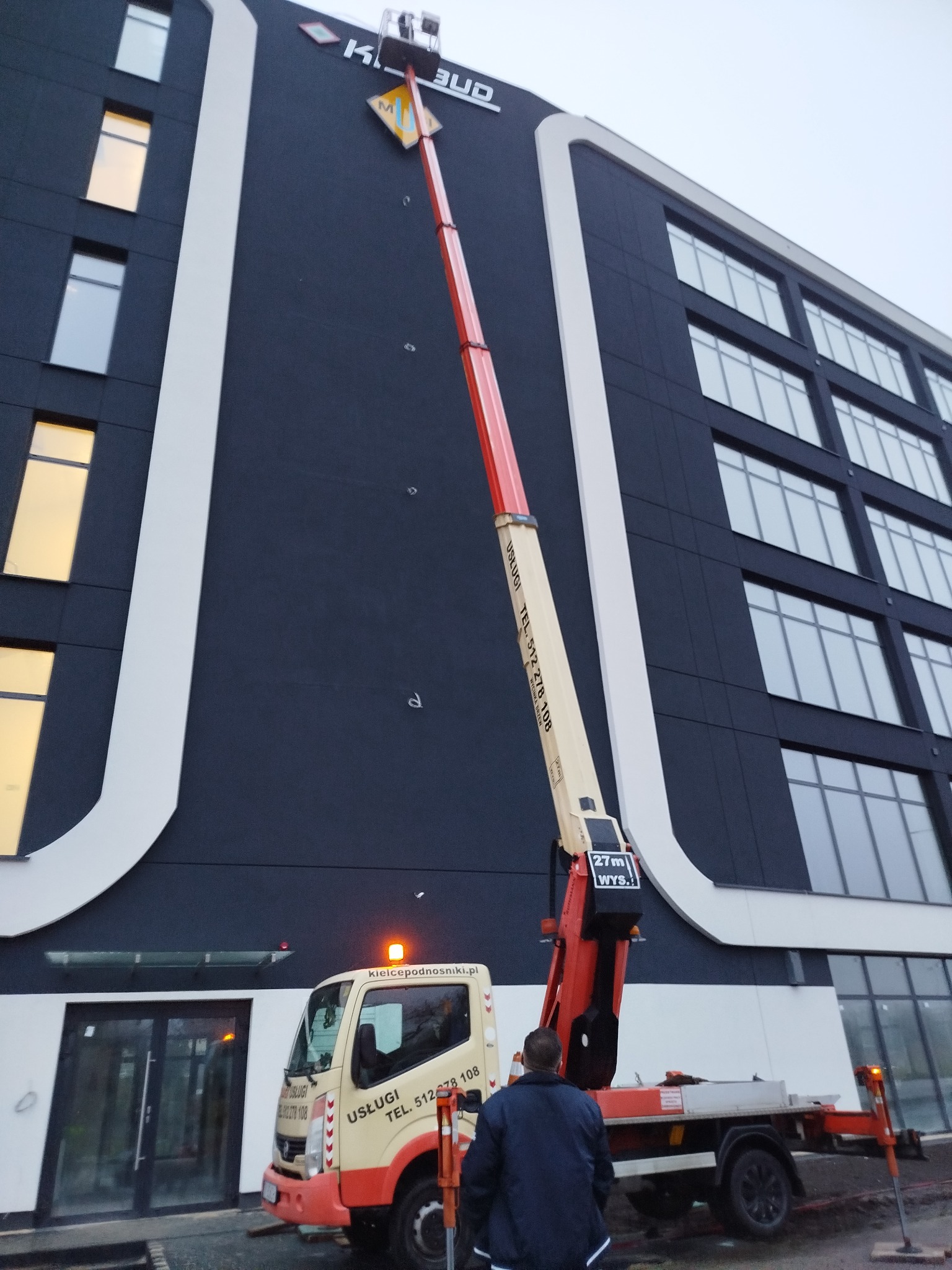 Podnośnik koszowy na tle nowoczesnego budynku biurowego z ciemną elewacją i białymi elementami architektonicznymi, widoczny pracownik w kurtce.