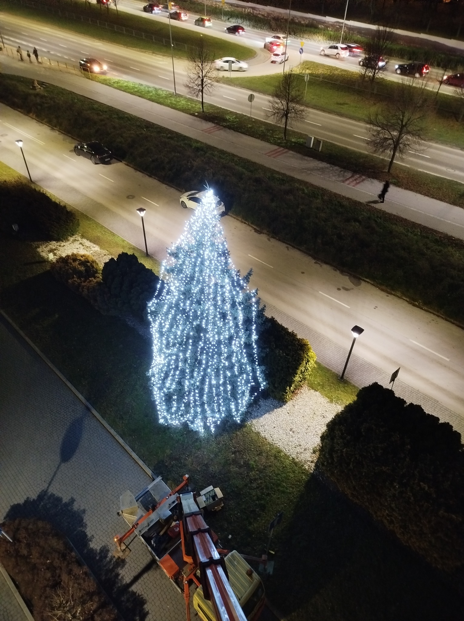 Widok z góry na oświetlone drzewko bożonarodzeniowe, ustawione w pasie zieleni przy ulicy, z podnośnika koszowego w trakcie montażu lub demontażu, nocne oświetlenie uliczne.