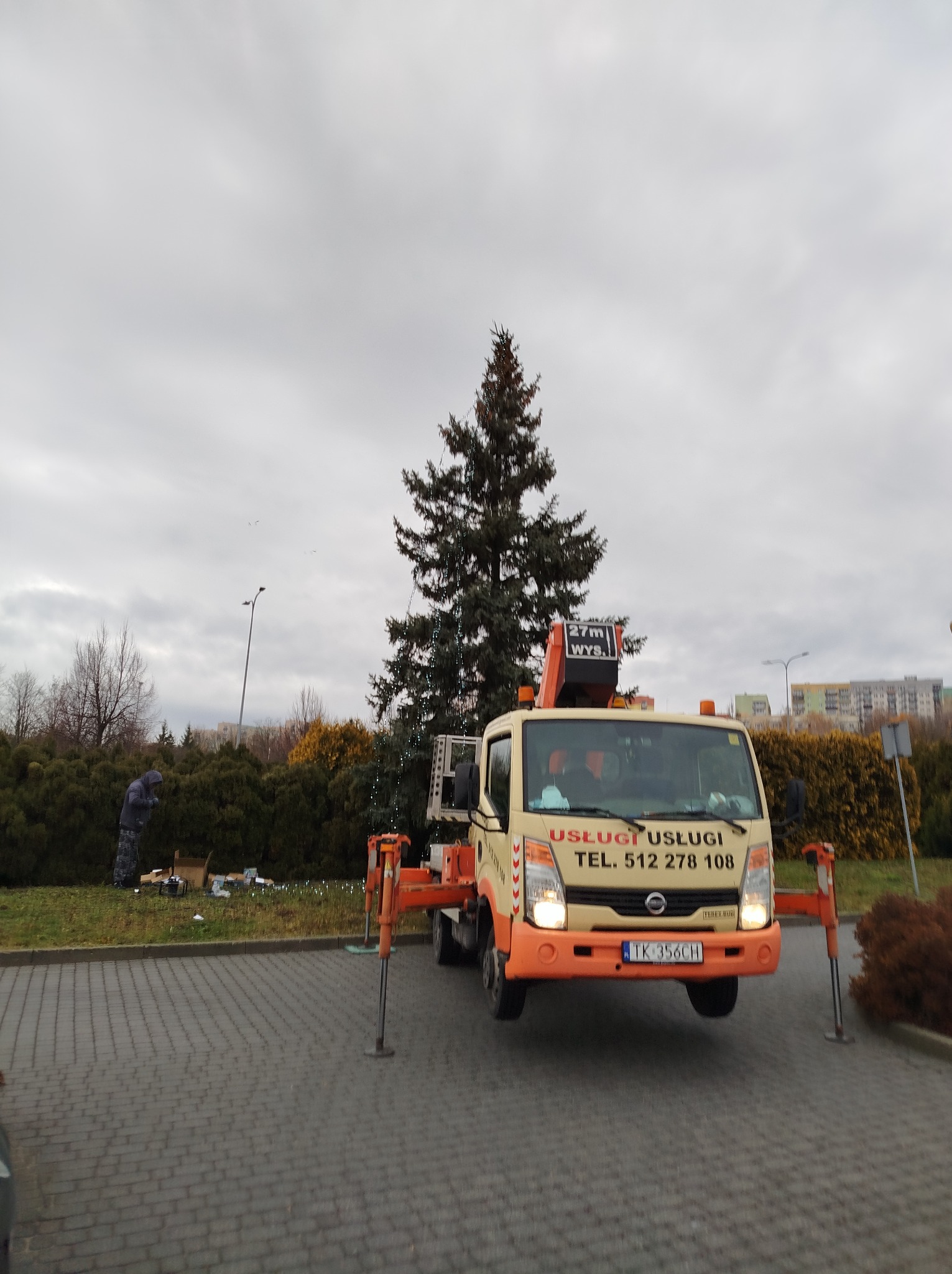 Żółto-pomarańczowy samochód z wysięgnikiem na parkingu, ozdobiona choinka w tle, pracownik w oddali przy dekoracjach.