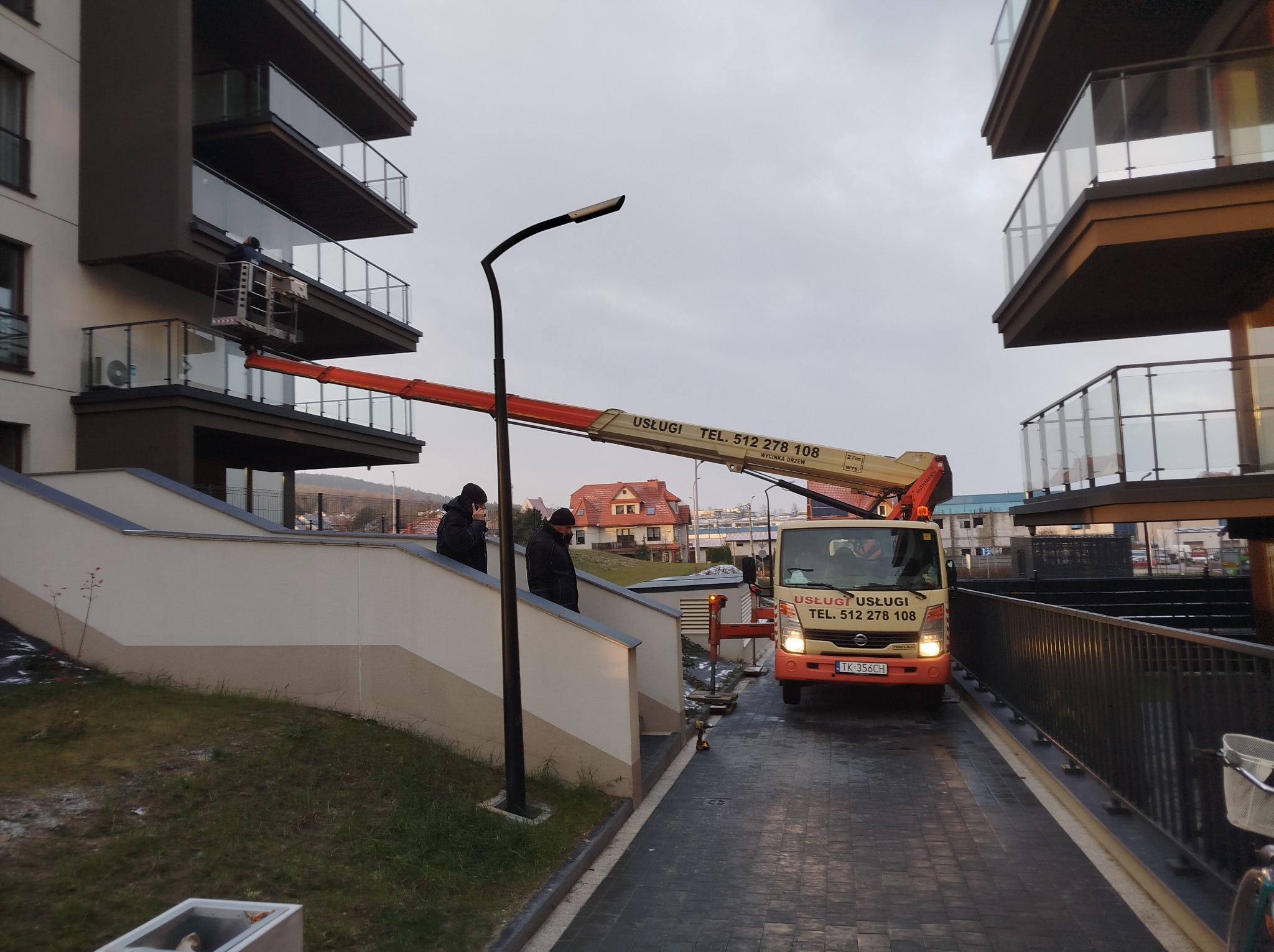 Pracownik na podnośniku koszowym przy balkonie nowoczesnego budynku, prawdopodobnie wykonujący prace pielęgnacyjne lub montażowe, z widoczną reklamą firmy na pojeździe oraz dwoma osobami...