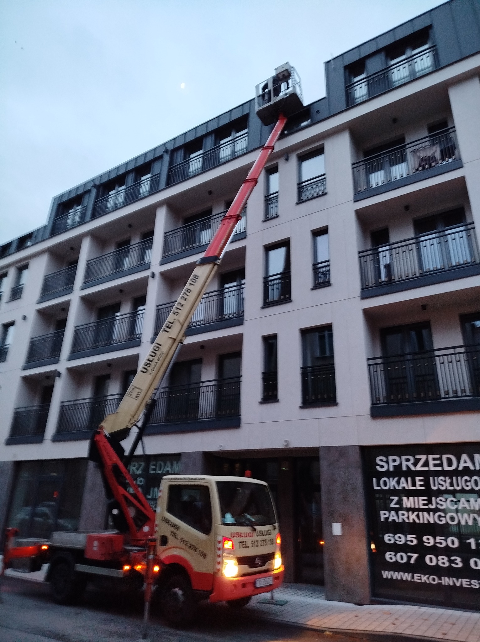 Podnośnik koszowy na tle nowoczesnego budynku mieszkalnego z balkonami; widoczny numer telefonu kontaktowego na ramieniu podnośnika.