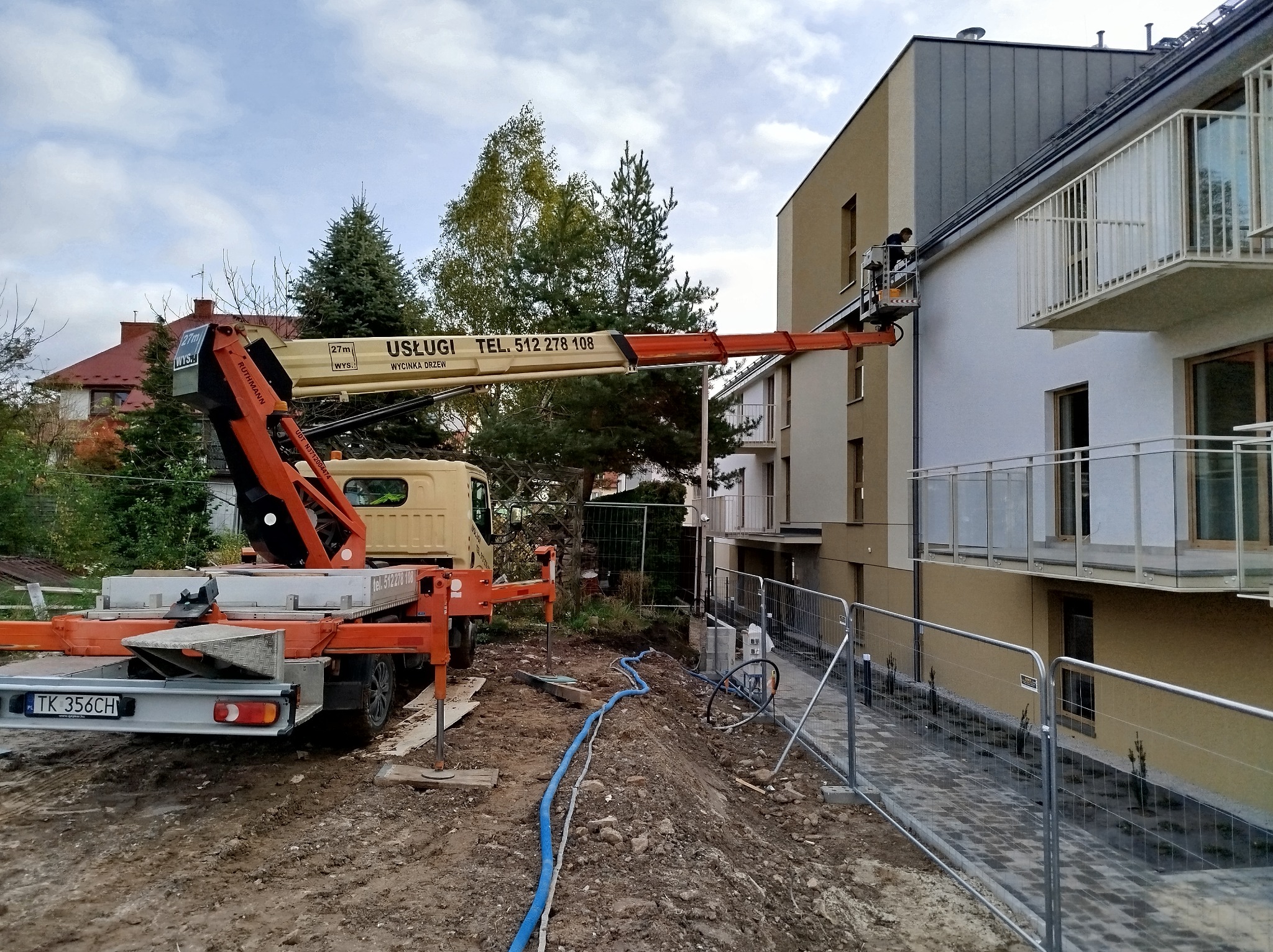 Pomarańczowy podnośnik koszowy na samochodzie ciężarowym, z napisem 'Usługi Wycinka Drzew' i numerem telefonu, ustawiony przy nowym budynku mieszkalnym; pracownik w koszu na wysokości okapu dachu.