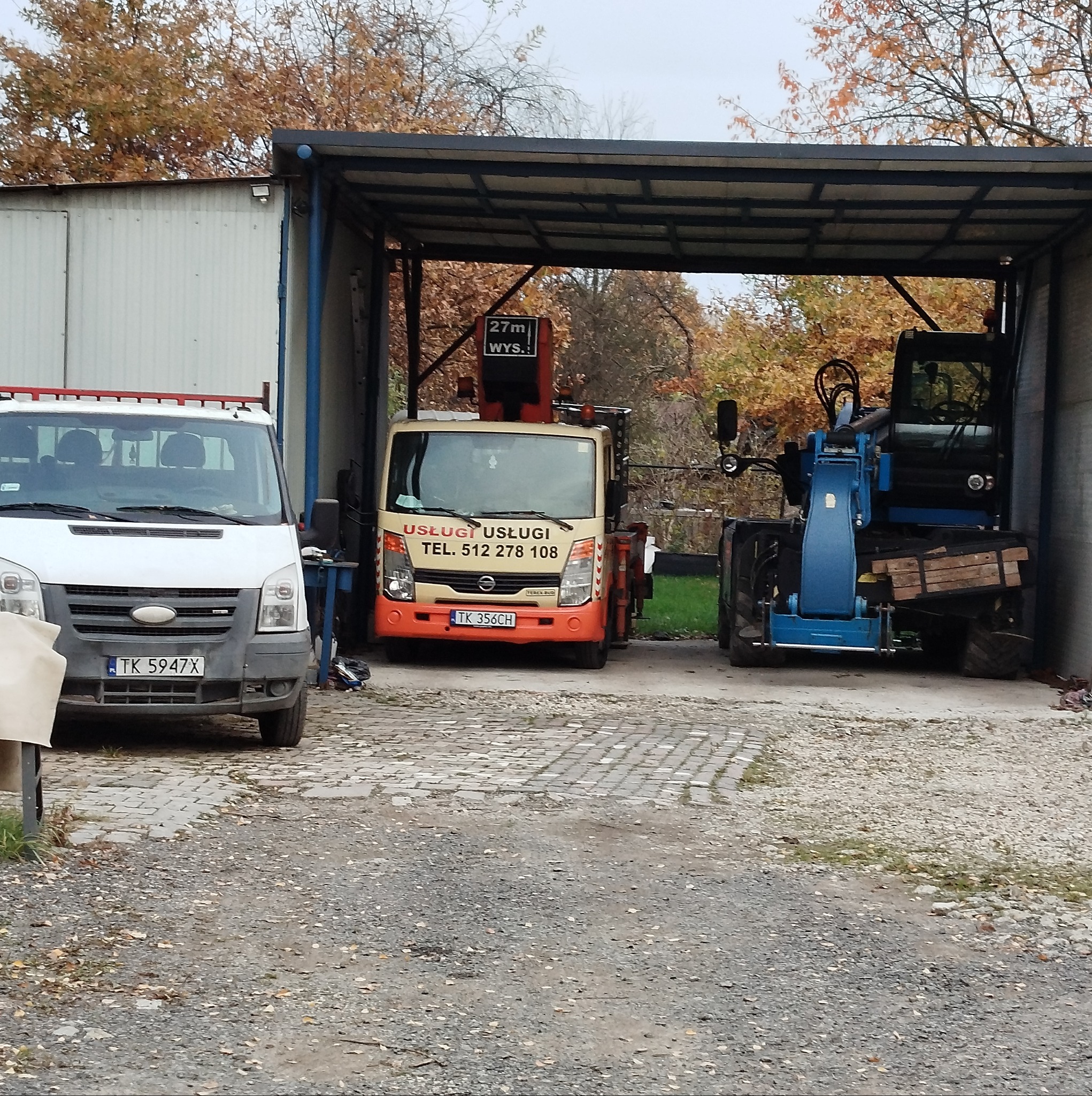 Flota pojazdów firmy: biały van, pomarańczowy samochód z dźwigiem oznaczony numerem telefonu i niebieska koparko-ładowarka, zaparkowane pod zadaszeniem na żwirowym placu, z jesiennymi drzewami w tle.