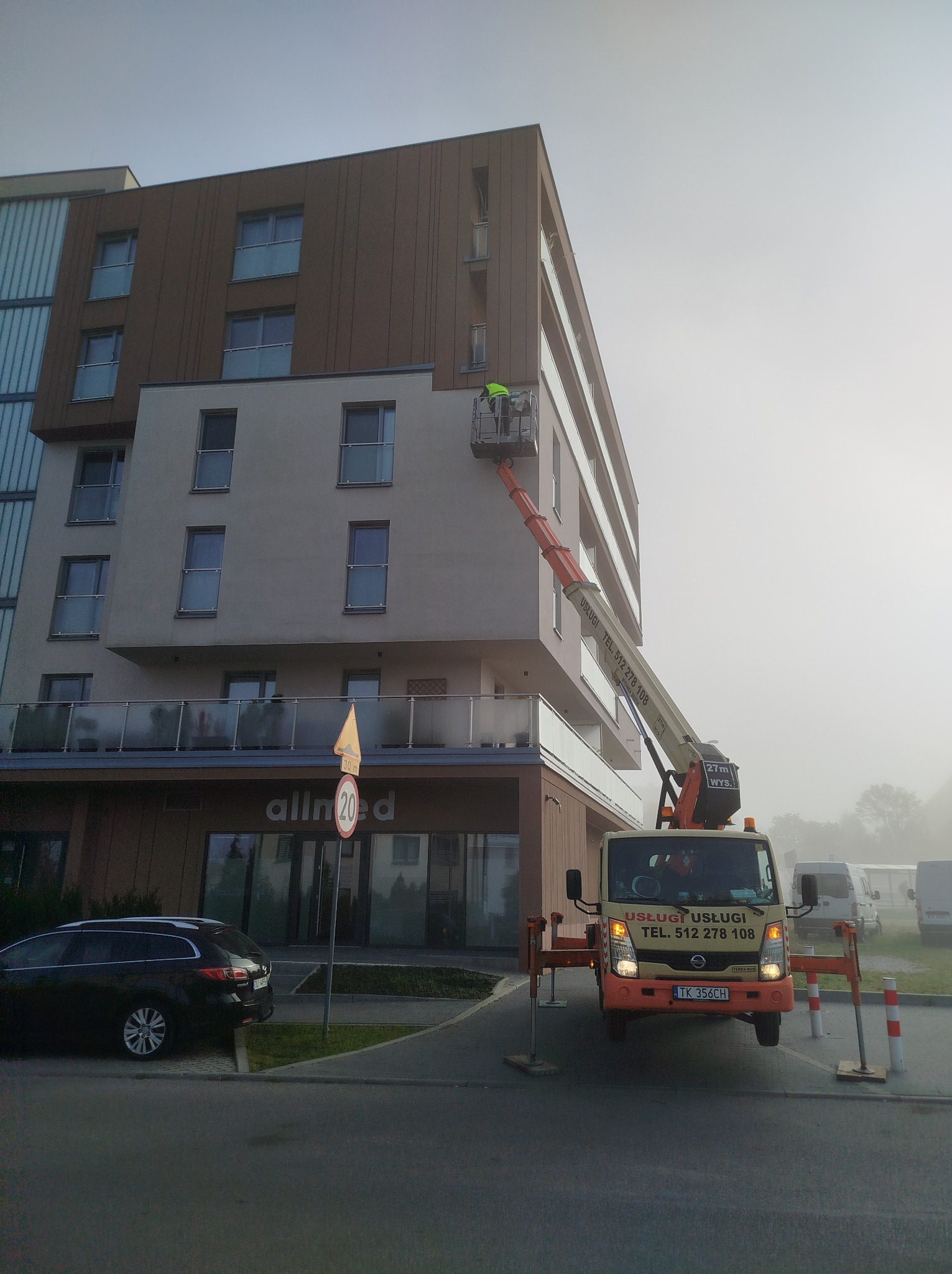 Pracownik w żółtej kamizelce na podnośniku koszowym przy elewacji budynku, na pierwszym planie zaparkowany samochód osobowy i oznakowanie drogowe, w tle zabudowa miejska w porannej mgle.