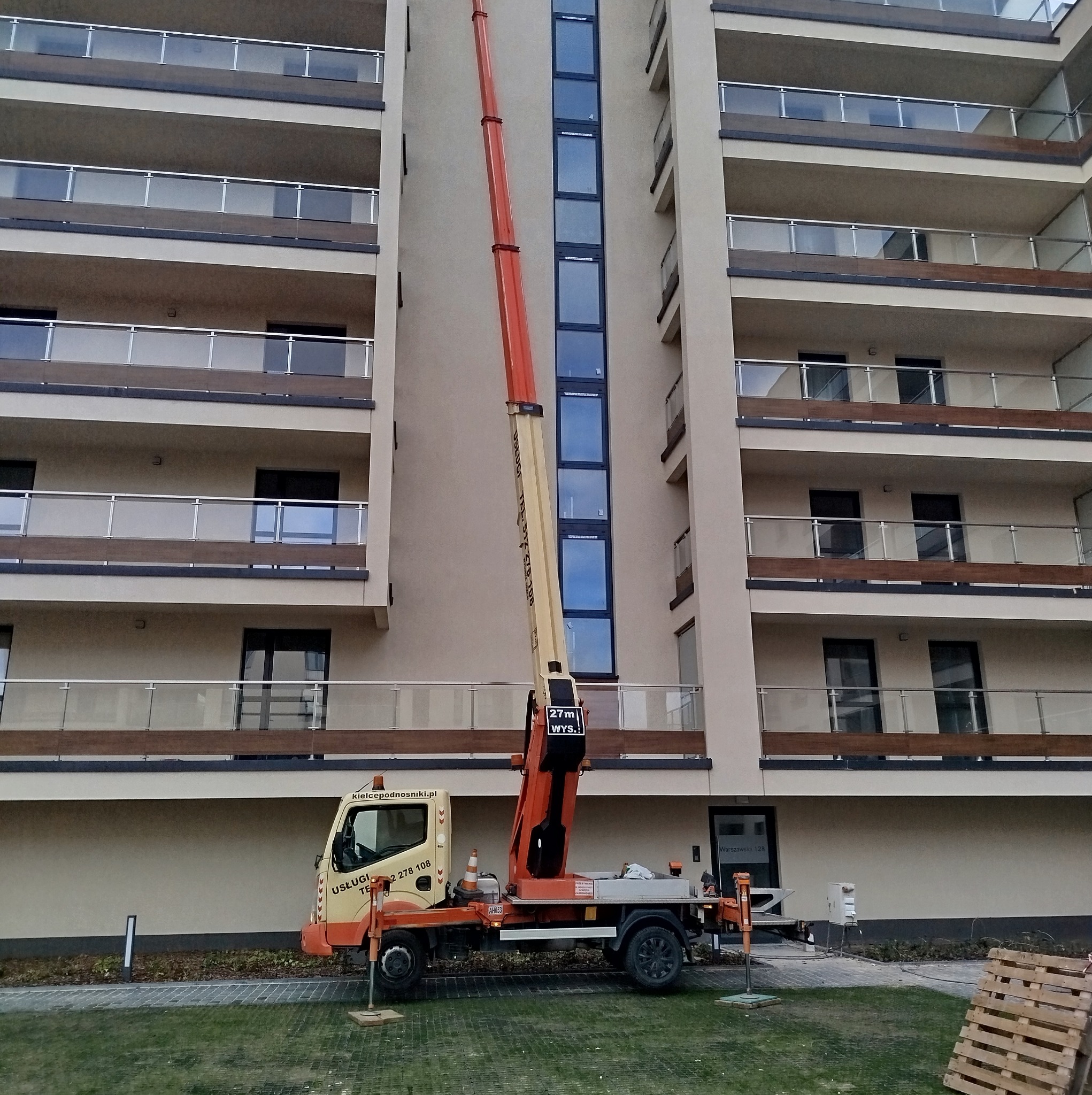 Pomarańczowo-beżowy wózek widłowy z wysięgnikiem ustawiony przed nowoczesnym budynkiem mieszkalnym z balkonami, przygotowany do prac na wysokości.