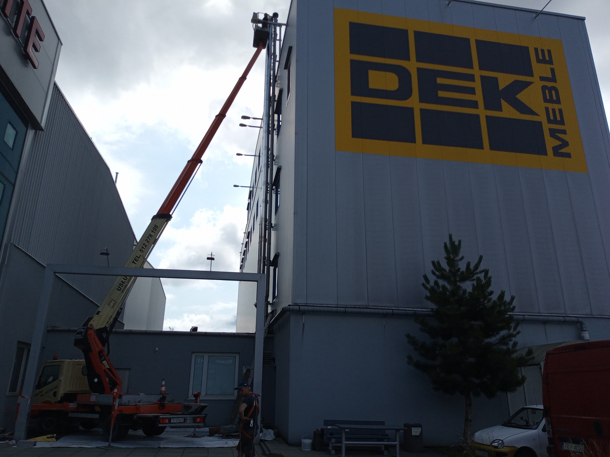 Praca na wysokościach przy budynku z użyciem podnośnika koszowego, widoczny pracownik w koszu i duży znak 'DEK MEBLE' na elewacji.
