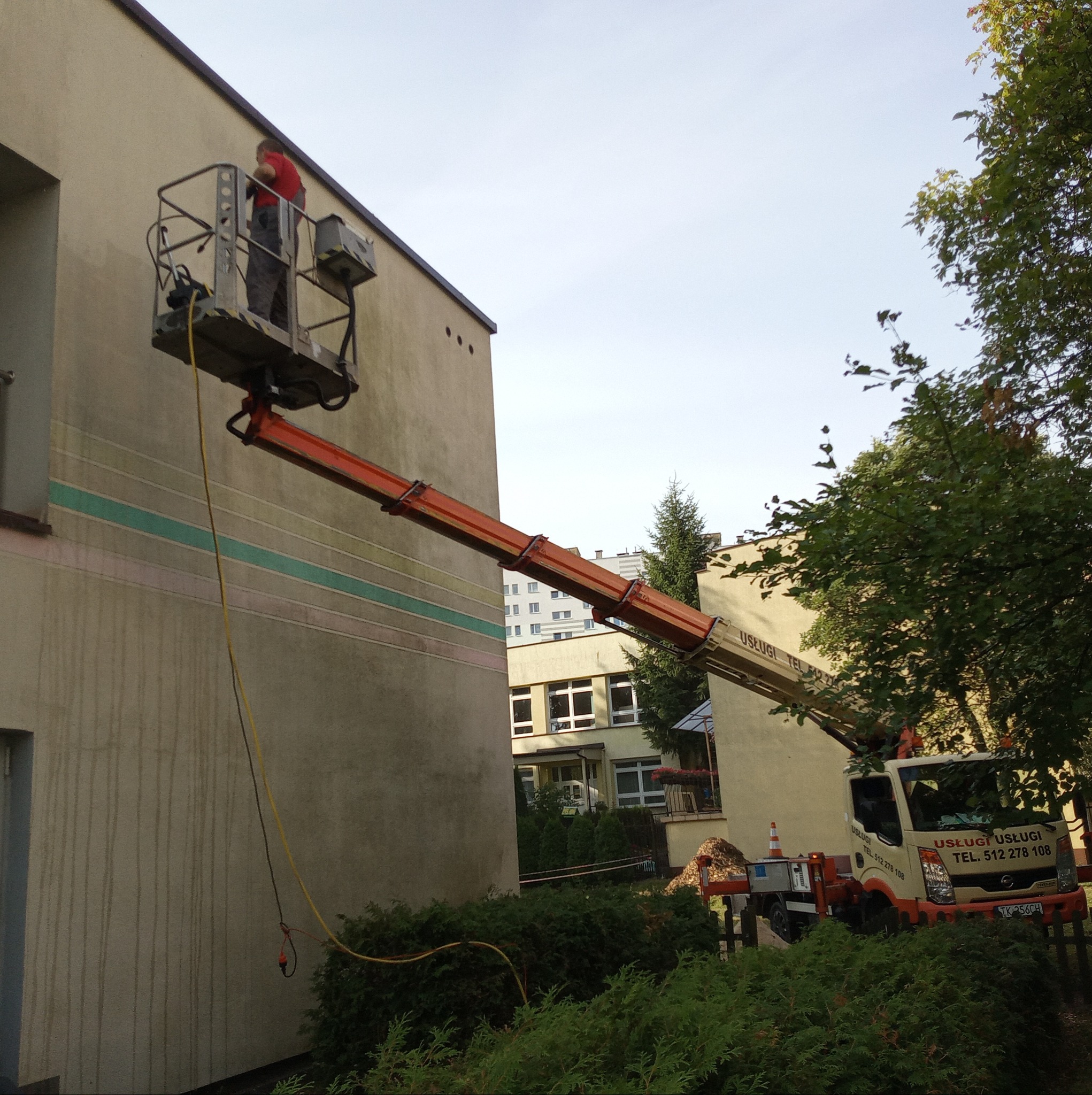Pracownik na podnośniku koszowym przy ścianie budynku, prawdopodobnie wykonujący prace konserwacyjne lub czyszczenie elewacji, z widocznym samochodem dostawczym i zielenią w tle.