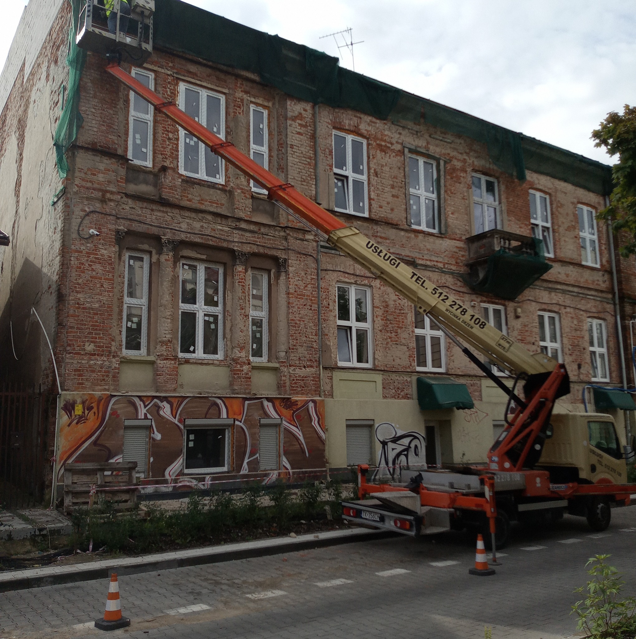 Podnośnik koszowy z pracownikiem na tle starej, ceglanej kamienicy z widocznymi śladami graffiti na dole elewacji i siatką zabezpieczającą na górze.