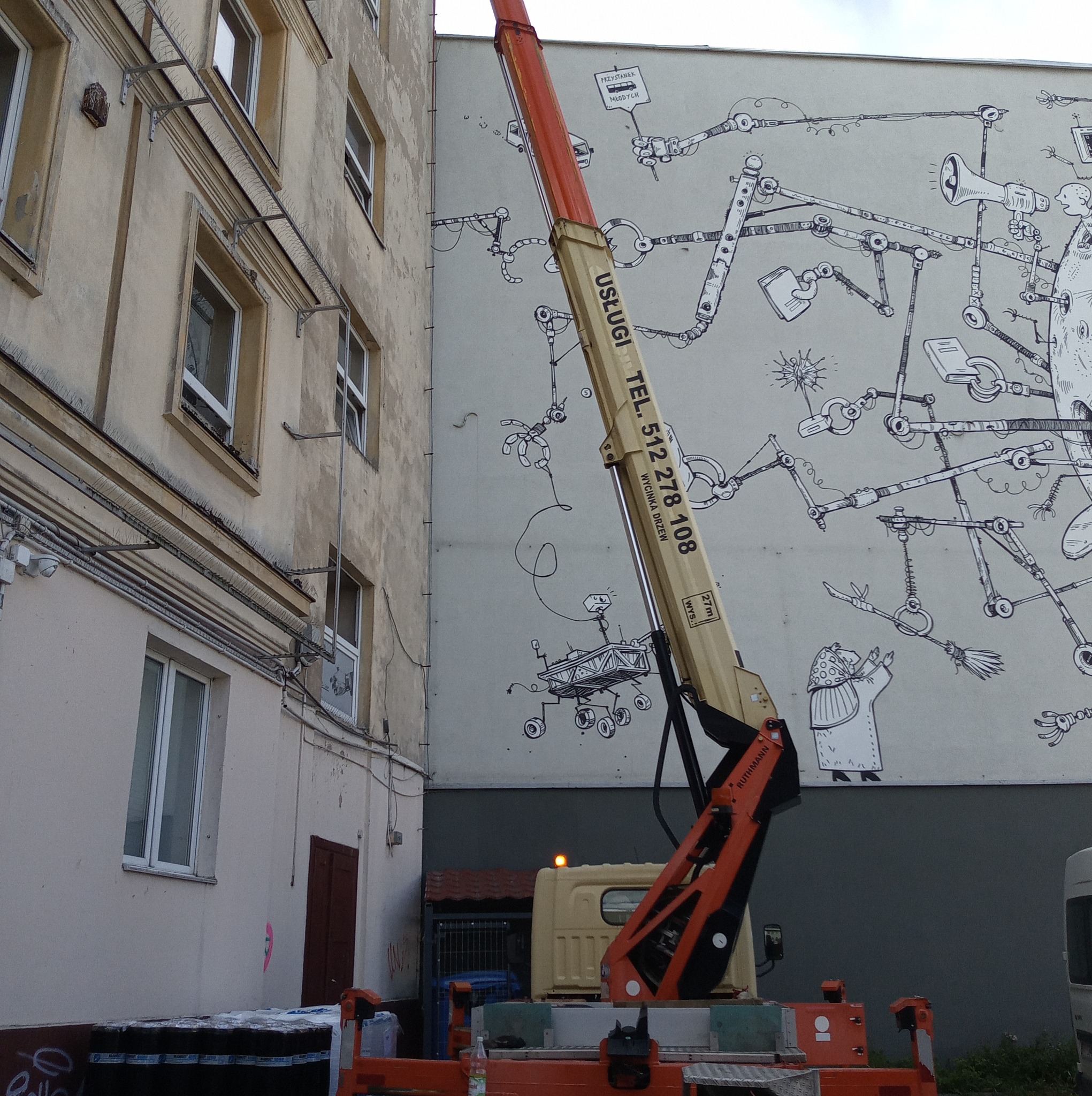 Wysięgnik z numerem telefonu na tle budynku z muralem w stylu science fiction, z widocznymi detalami konstrukcyjnymi i elementami otoczenia.