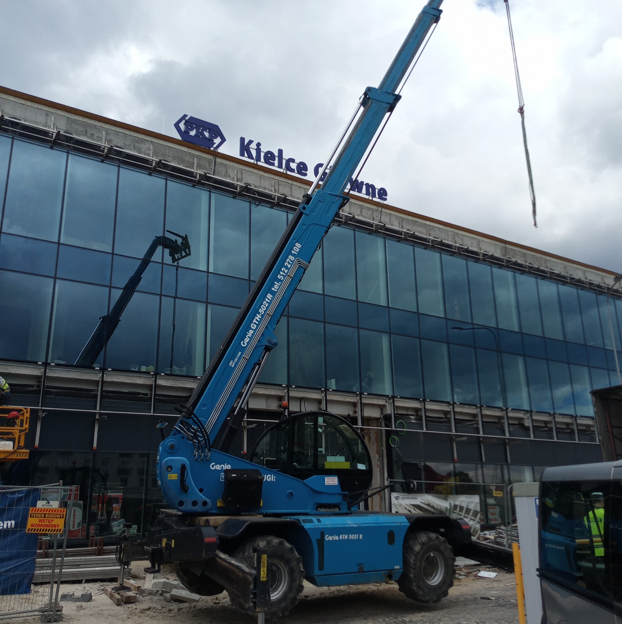 Niebieski wózek widłowy teleskopowy Genie GTH-5021R na placu budowy przed budynkiem z napisem 'Kielce Główne' i logiem PKP na dachu, widoczne rusztowania i materiały budowlane.