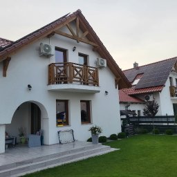 Biały dom z drewnianymi elementami i balkonem, na ścianie zamontowane klimatyzatory. Drugi dom w tle z panelami słonecznymi na dachu. Zadbany trawnik.