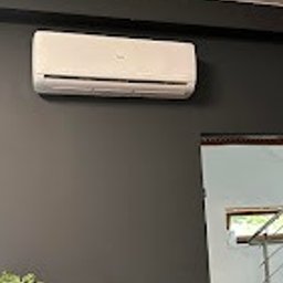 Biały klimatyzator ścienny na ciemnoszarej ścianie, widoczny fragment schodów i rośliny doniczkowej. Minimalistyczny design wnętrza z nowoczesnym urządzeniem chłodzącym.