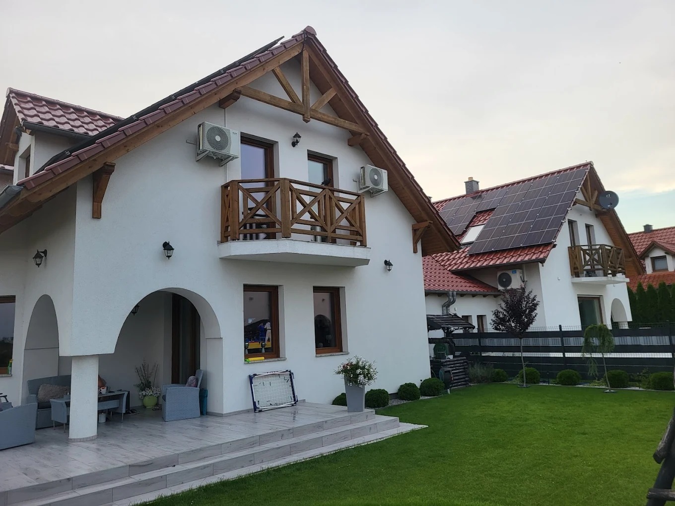 Biały dom z drewnianymi elementami i balkonem, na ścianie zamontowane klimatyzatory. Drugi dom w tle z panelami słonecznymi na dachu. Zadbany trawnik.