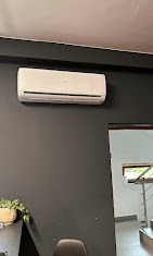 Biały klimatyzator ścienny na ciemnoszarej ścianie, widoczny fragment schodów i rośliny doniczkowej. Minimalistyczny design wnętrza z nowoczesnym urządzeniem chłodzącym.