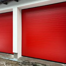 Dwie czerwone, segmentowe bramy garażowe w białej elewacji budynku. Widoczne kostki brukowe przed jedną z bram. Na ścianie budynku naklejona informacja o budowie.