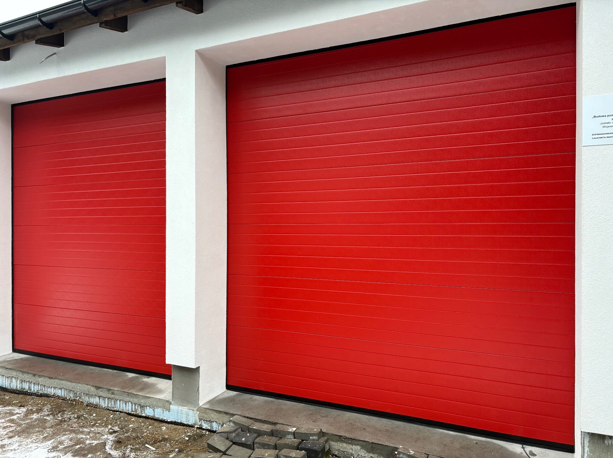 Dwie czerwone, segmentowe bramy garażowe w białej elewacji budynku. Widoczne kostki brukowe przed jedną z bram. Na ścianie budynku naklejona informacja o budowie.