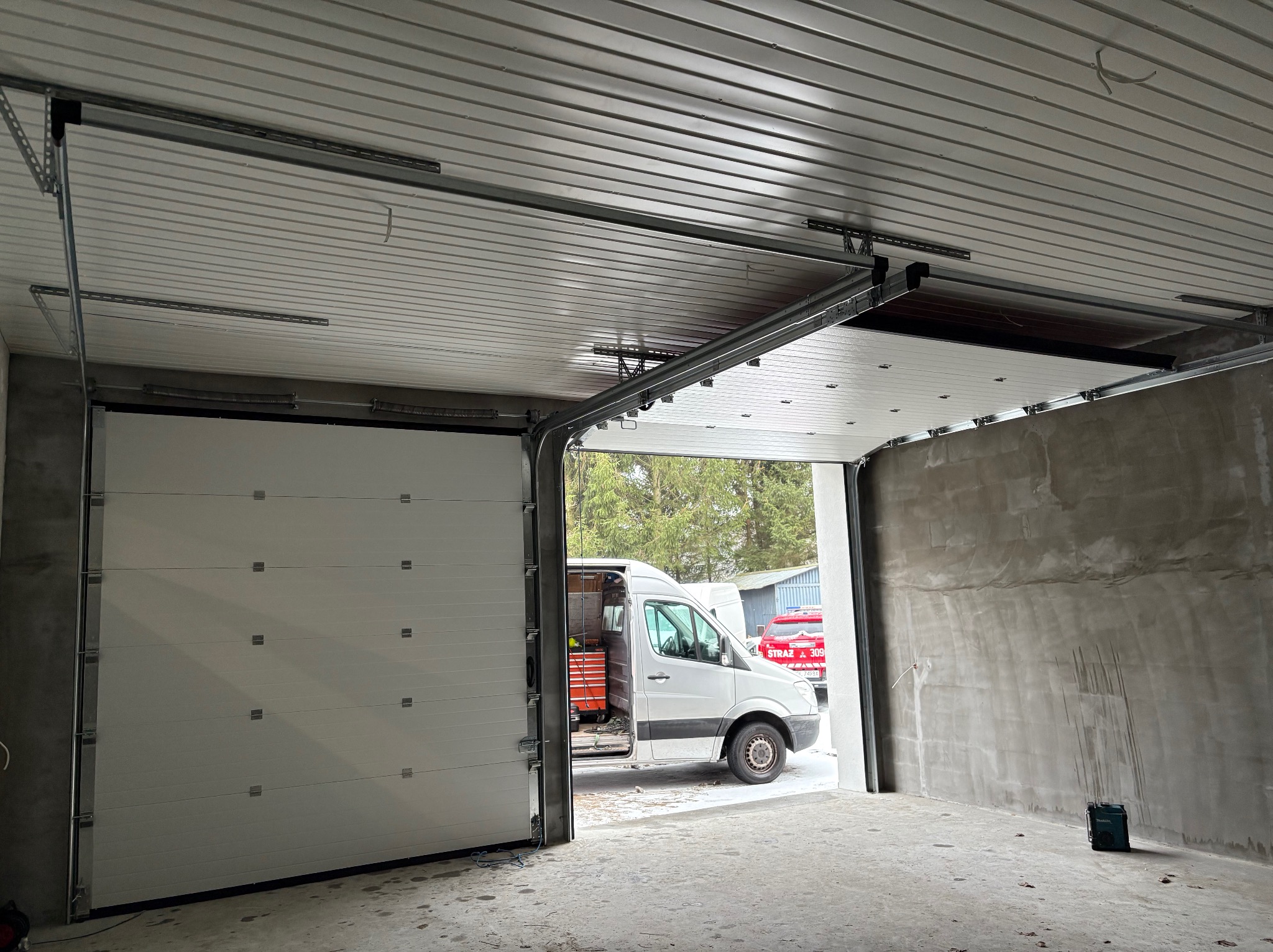 Częściowo otwarta segmentowa brama garażowa z widocznymi prowadnicami pod sufitem, na zewnątrz zaparkowany biały bus z otwartymi drzwiami i skrzynką narzędziową w środku, surowe ściany garażu w tle.