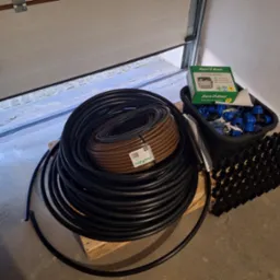 Zestaw elementów do instalacji nawadniania: zwój czarnego węża nawadniającego, brązowy wąż kroplujący, sterownik Rain Bird ESP-TM2, złączki i kratka trawnikowa, ułożone na palecie w garażu.